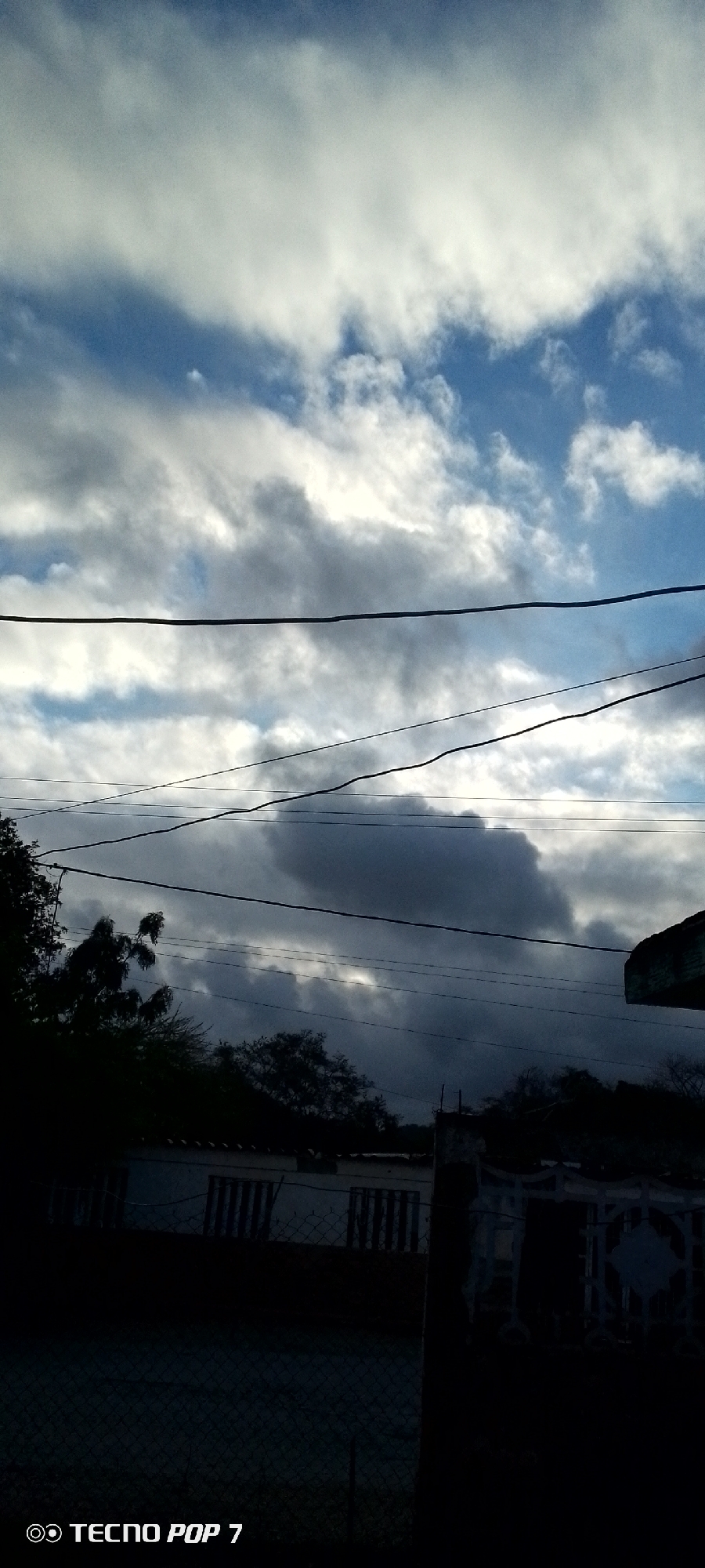 The image depicts a view of a sky filled with a mix of white and dark clouds, with patches of blue sky visible. Several power lines are stretched horizontally across the frame. In the foreground, silhouetted against the sky, are the tops of trees and the roofline of a building, with what appears to be a fence in front of it. The lower portion of the image is very dark, obscuring details of the ground and any potential inhabitants. A watermark at the bottom left reads "TECNO POP 7". The scene is consistent with an outdoor view in Altagracia, Venezuela, characterized by residential structures and overhead electrical infrastructure against a dynamic cloudscape.