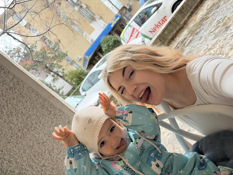The image displays an adult female and a young child in an outdoor urban setting within Budva, Montenegro. The adult female, positioned on the right, possesses blonde hair and is depicted winking with her tongue extended. She is wearing a light-colored, long-sleeved top. To her left, a young child wears a light blue full-body suit adorned with castle and animal patterns, and a light beige knitted hat. The child is also extending its tongue and has both hands raised to the sides of its head in a playful gesture. The two individuals are in close proximity, suggesting an interaction or shared moment. In the background, a street scene is visible, featuring several parked vehicles, including a white delivery van with prominent "Nektar" branding on its side. Bare-branched trees and multi-story buildings with balconies are present, one featuring a blue awning and a partially visible "Goodyear" sign. A speckled, possibly granite, wall texture is seen on the left side of the frame, and a reflective surface, possibly wet ground or a table, occupies the lower right.