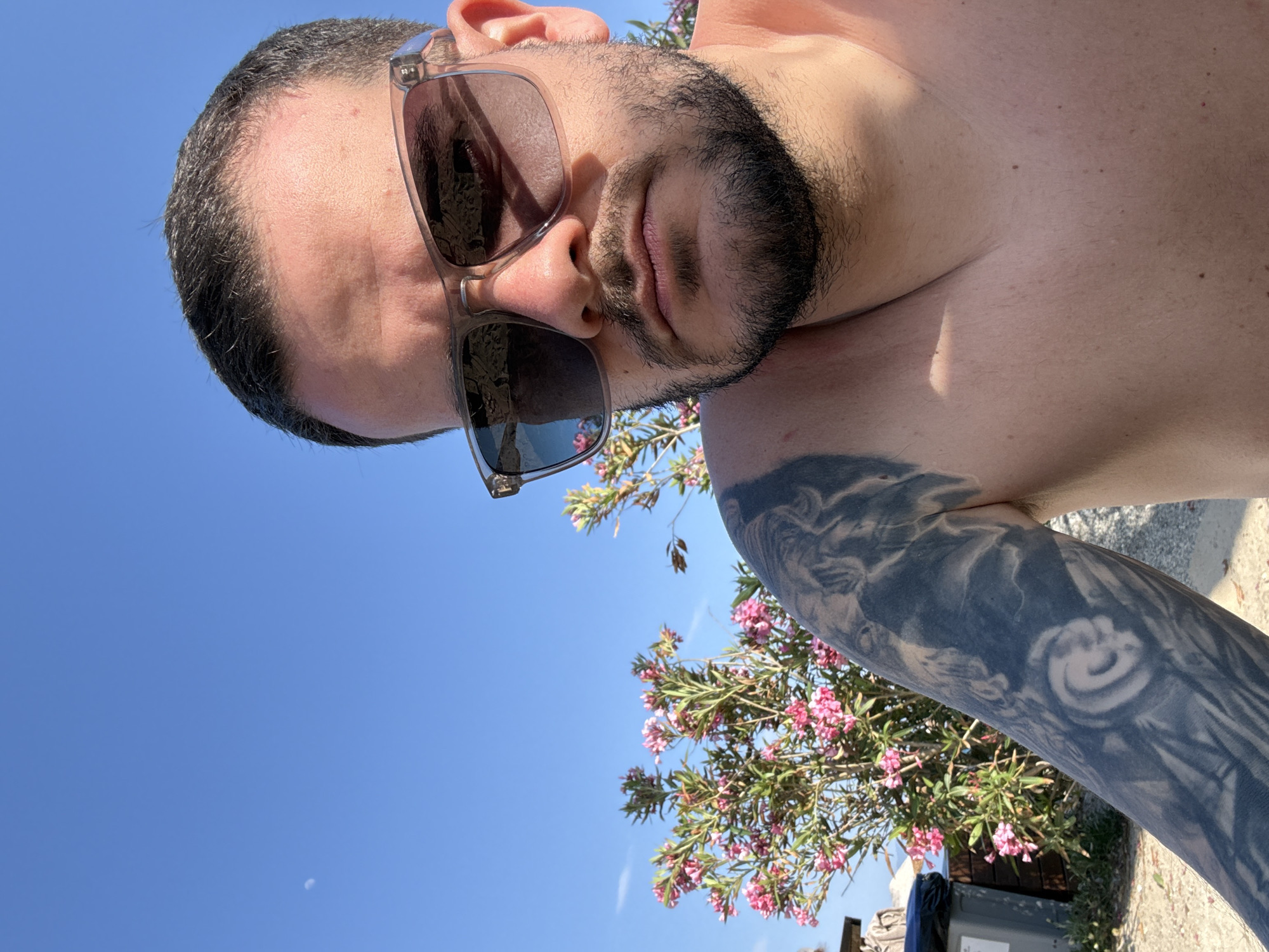 A close-up, tilted selfie shows a man with a beard, wearing sunglasses. He has short, dark hair and a tattoo on his left arm. Behind him, there are pink flowers and a clear blue sky. The location is likely Kos, Greece, as this information was provided by the user. A blue trash bin is visible in the lower right corner. The angle suggests the photo was taken from a low vantage point, looking up at the man.