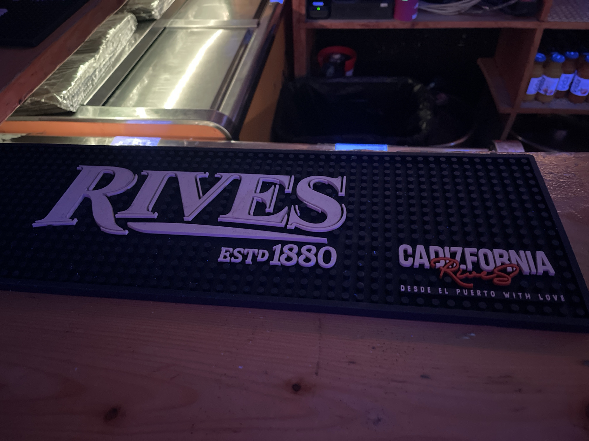 The image shows a close-up view of a black bar mat with embossed white text. The text on the left reads "RIVES ESTD 1880". On the right, the text reads "CADIZFORNIA" with a stylized red cursive logo in the middle and "DESDE EL PUERTO WITH LOVE" below it. The bar mat is positioned on a wooden surface, likely a bar counter. In the background, out of focus, are stainless steel surfaces, some bottles on shelves, and dark cabinets. There are no people visible in the image. The lighting suggests an indoor environment, possibly a bar or restaurant. The mention of Barcelona, Spain, is not supported by any visual evidence within the image itself.