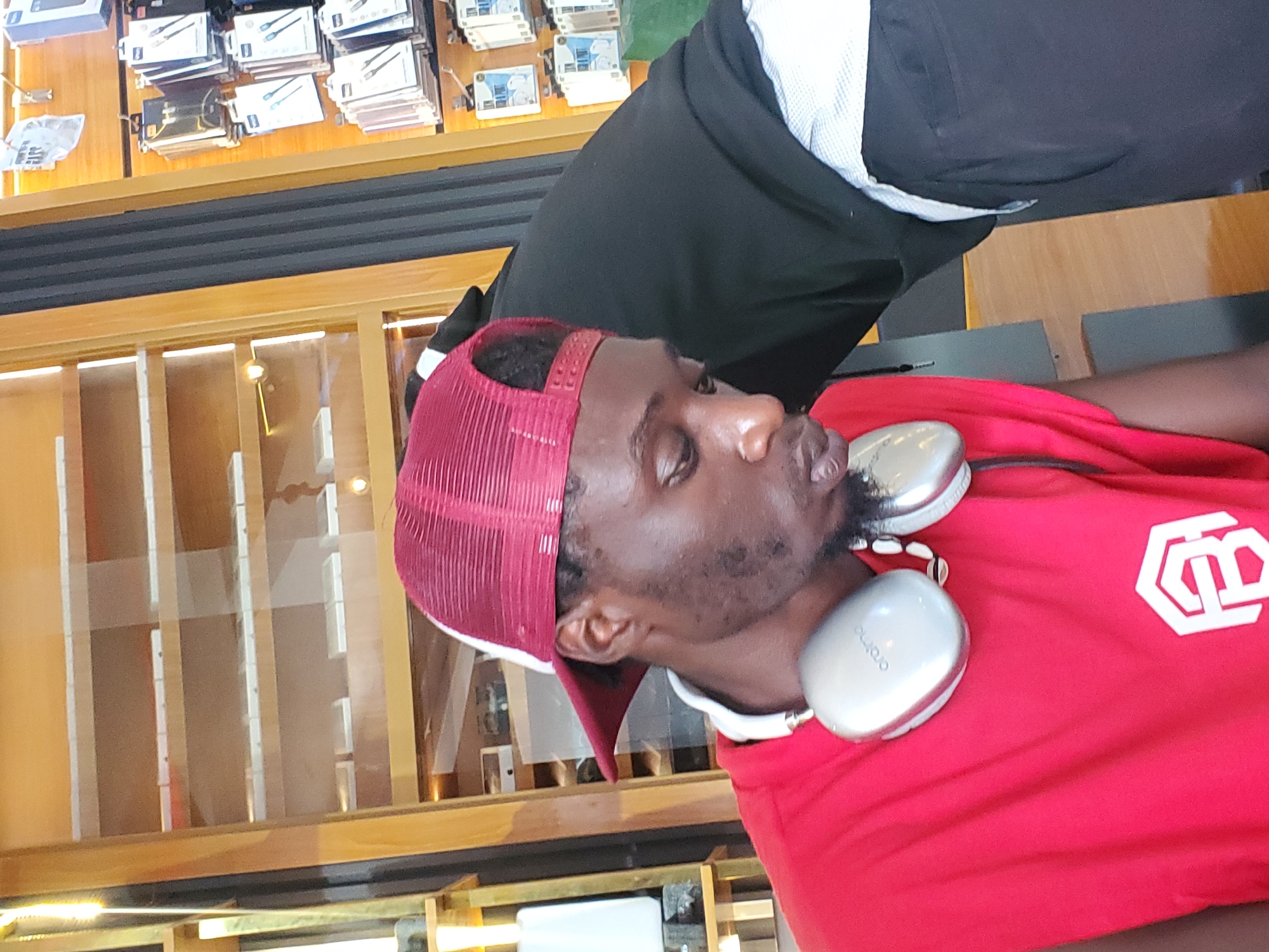 A man is wearing a red trucker hat and a red t-shirt with a white logo on it. He has white headphones on. He appears to be in a store with shelves stocked with small boxed items, possibly electronics accessories like phone chargers. The lighting suggests it could be daytime, as the store appears well-lit. The man's expression is neutral. The text "oraimo" is visible on one of the headphone earcups. The image is oriented vertically.
