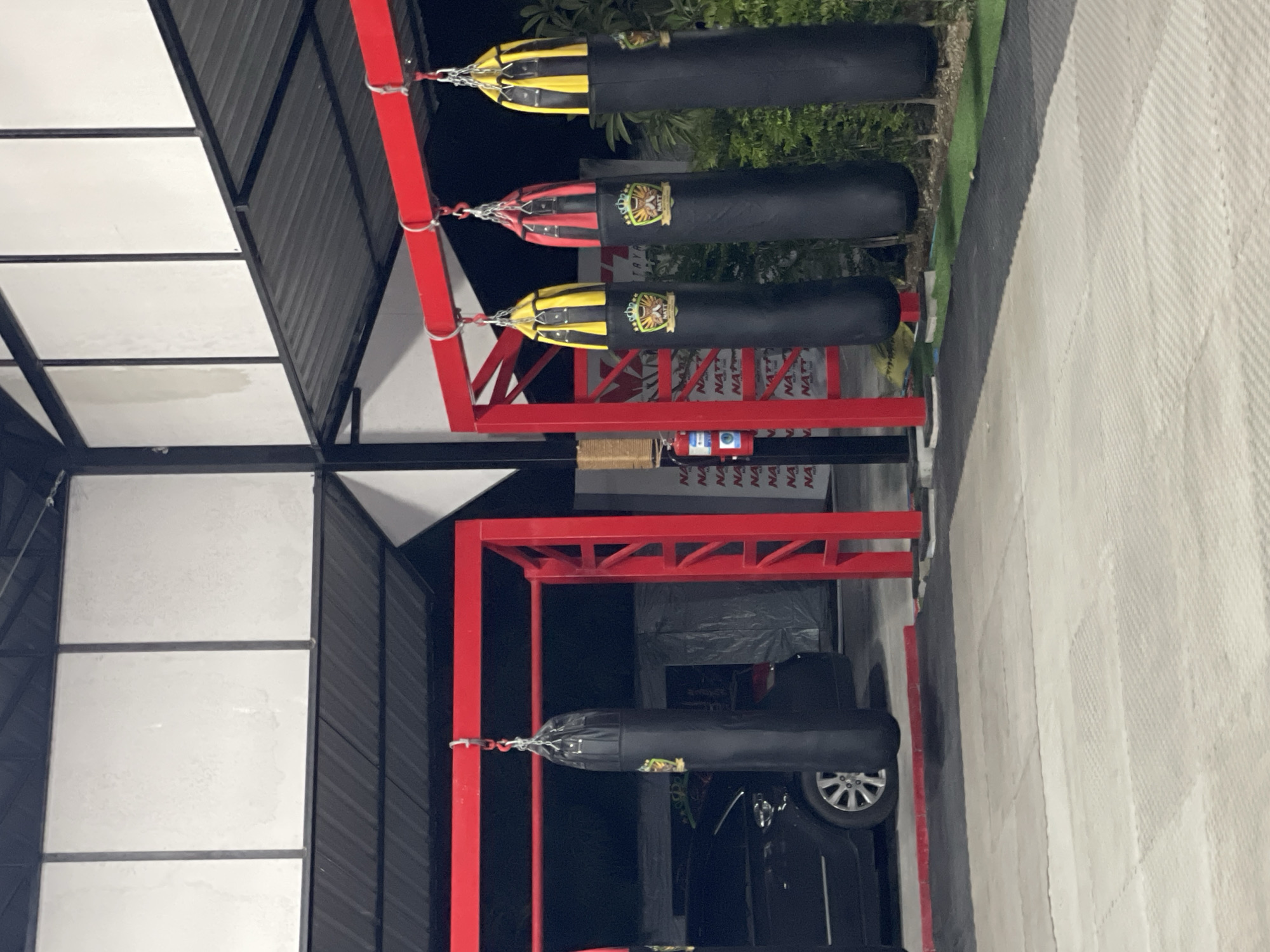 In Pattaya City, Thailand, an outdoor Muay Thai training facility features a red metal rack from which four black punching bags are suspended. The bags have yellow and red tops and display crests with text. In the background, a black car is parked. The ground is covered with interlocking mats, and a green artificial turf area is visible to the right. A fire extinguisher is mounted on a pole to the left of the bags. The facility appears to be covered by a corrugated metal roof.