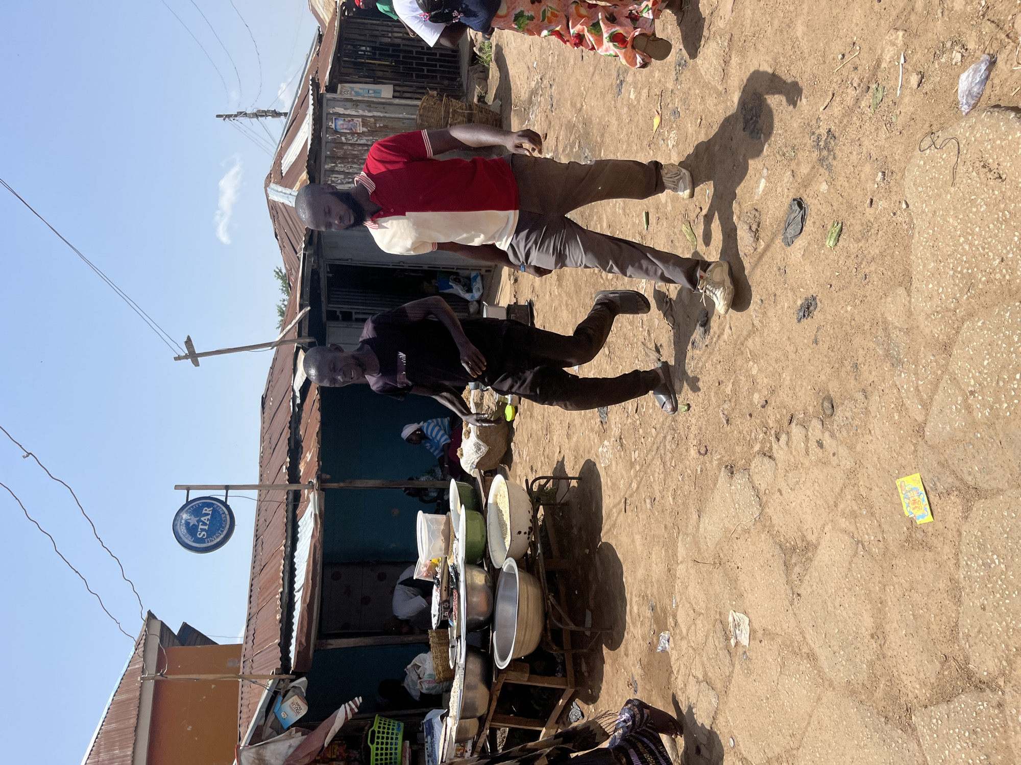 Two men walk in a street market. One man on the left wears a black t-shirt and black pants. The man on the right wears a red and white half-and-half polo shirt and gray pants. In the background, there are market stalls with various goods displayed in bowls and baskets. A blue sign with the word "STAR" in white lettering is visible above one of the stalls. The ground is dusty and uneven, with some debris scattered around. The scene appears to be in Gwong, Nigeria, with the architecture and market setup being consistent with the region. The sky is clear and blue, indicating a sunny day.