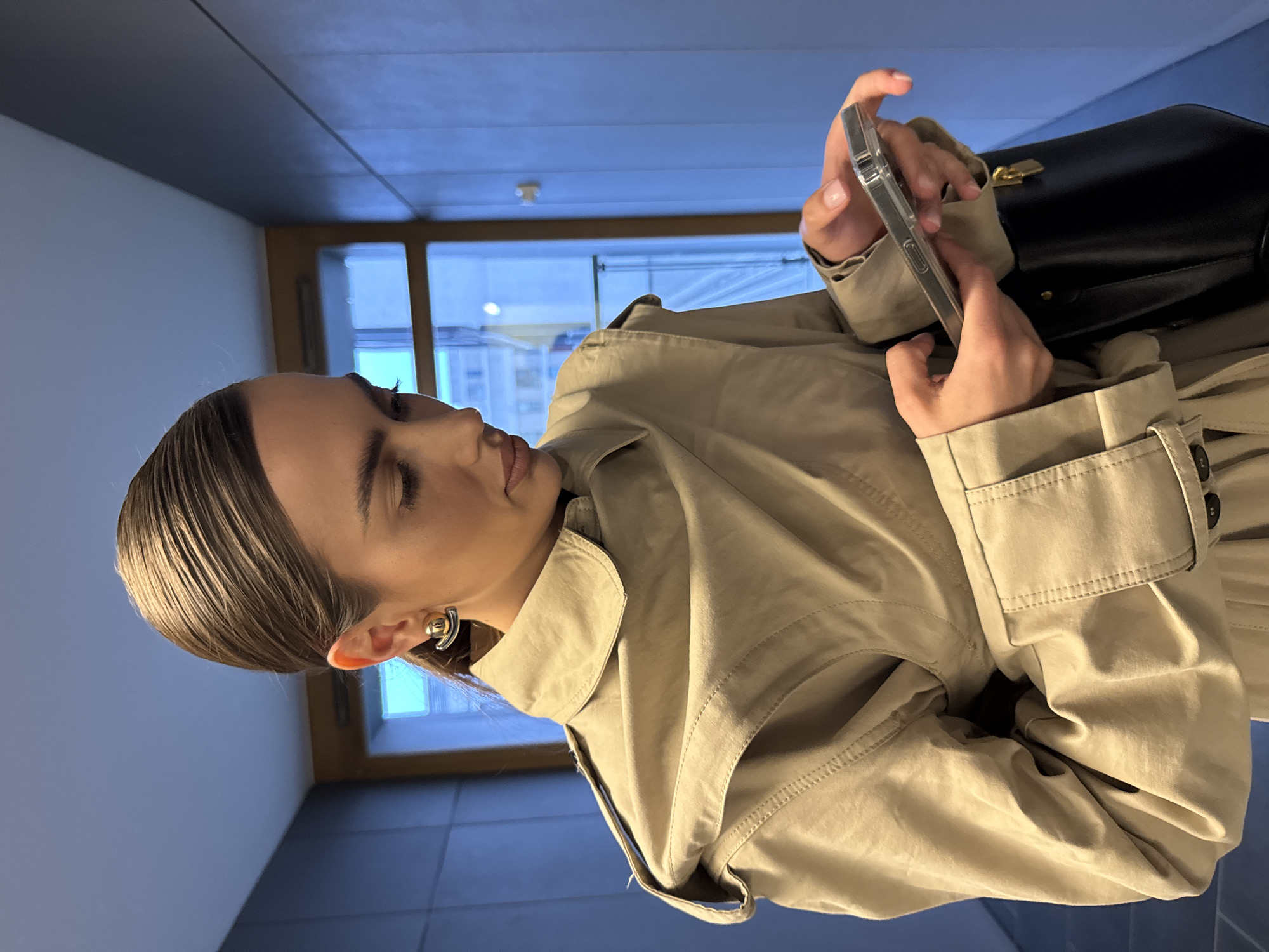 A woman is standing indoors in a building with blue walls. She is wearing a beige trench coat and holding a cell phone in her hands. A black purse hangs nearby. She has short, slicked-back hair. There is a window with white frames behind her. The exact location in the city of Unknown, Unknown is indeterminable from the image.