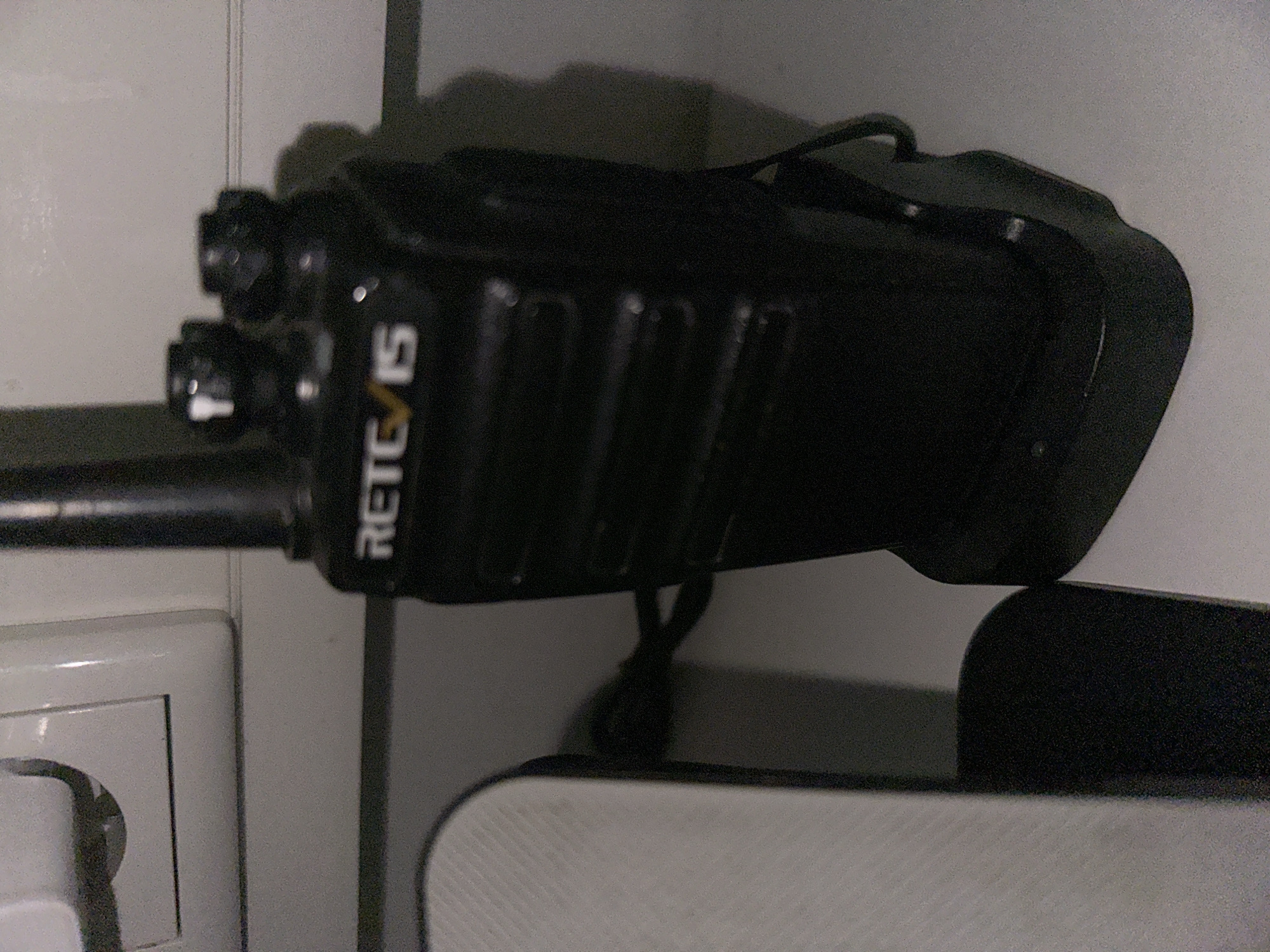 A black two-way radio charger is on a white surface next to a white electrical outlet. The charger is partially obscured by a white object on the left. The radio is standing upright in the charger. The charger has a black cord connected to it. The background is a plain white wall.