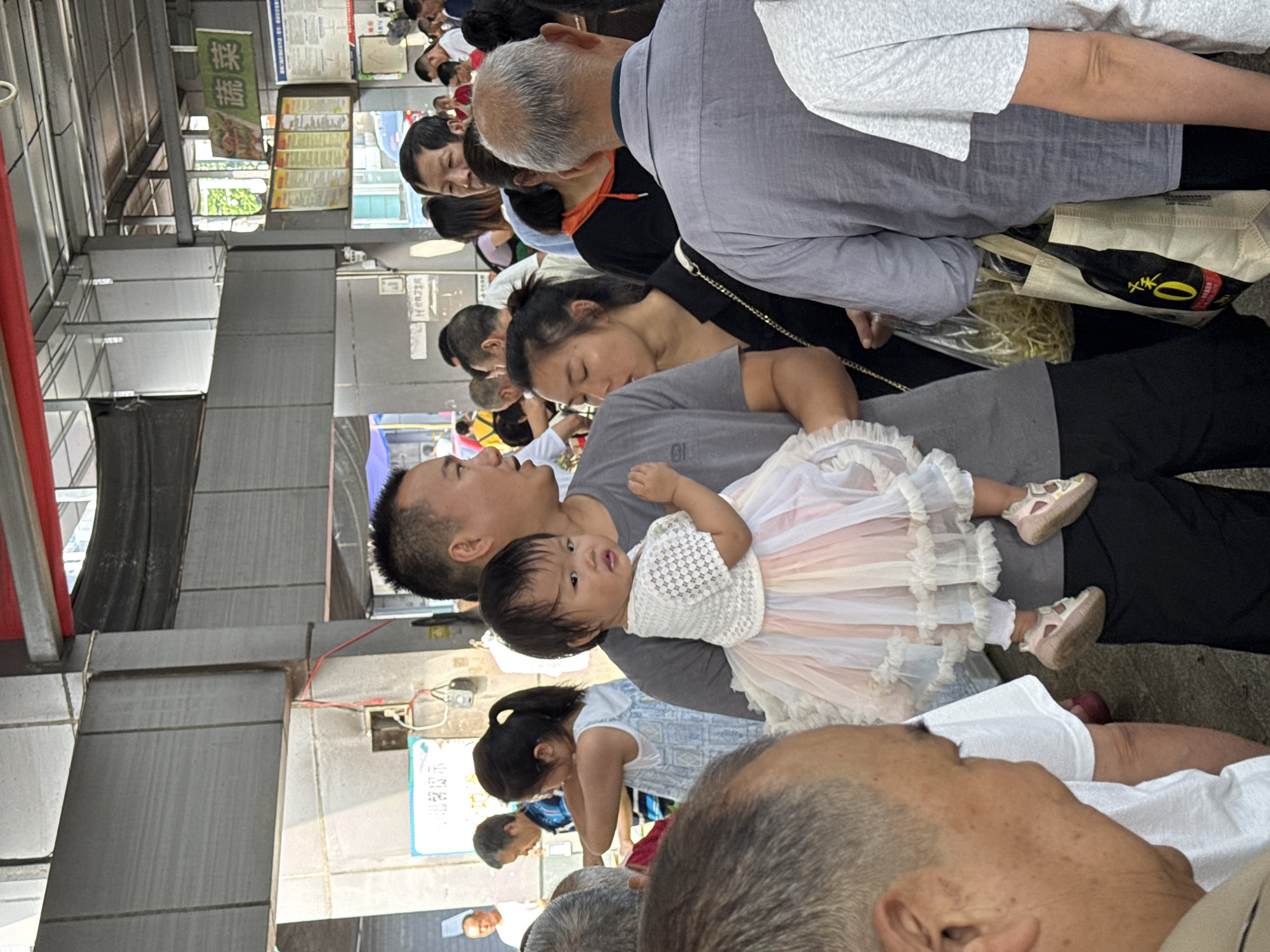 The image depicts a crowded scene, likely a market or public gathering space, in Jiangjin, China. In the foreground, a man is holding a young child wearing a frilly dress and pink shoes. The man is looking to the right, while the child is looking towards the viewer. Behind them, a woman in a black top with a gold chain strap is visible, appearing to be engaged in conversation or observing the surroundings. Further back, a crowd of people of various ages are present, some walking, some standing, and some interacting with each other. The environment appears to be an open-air or covered market, with structural elements like concrete columns and metal beams visible. Signage in Chinese characters can be seen on the buildings in the background. The lighting suggests it is daytime.
