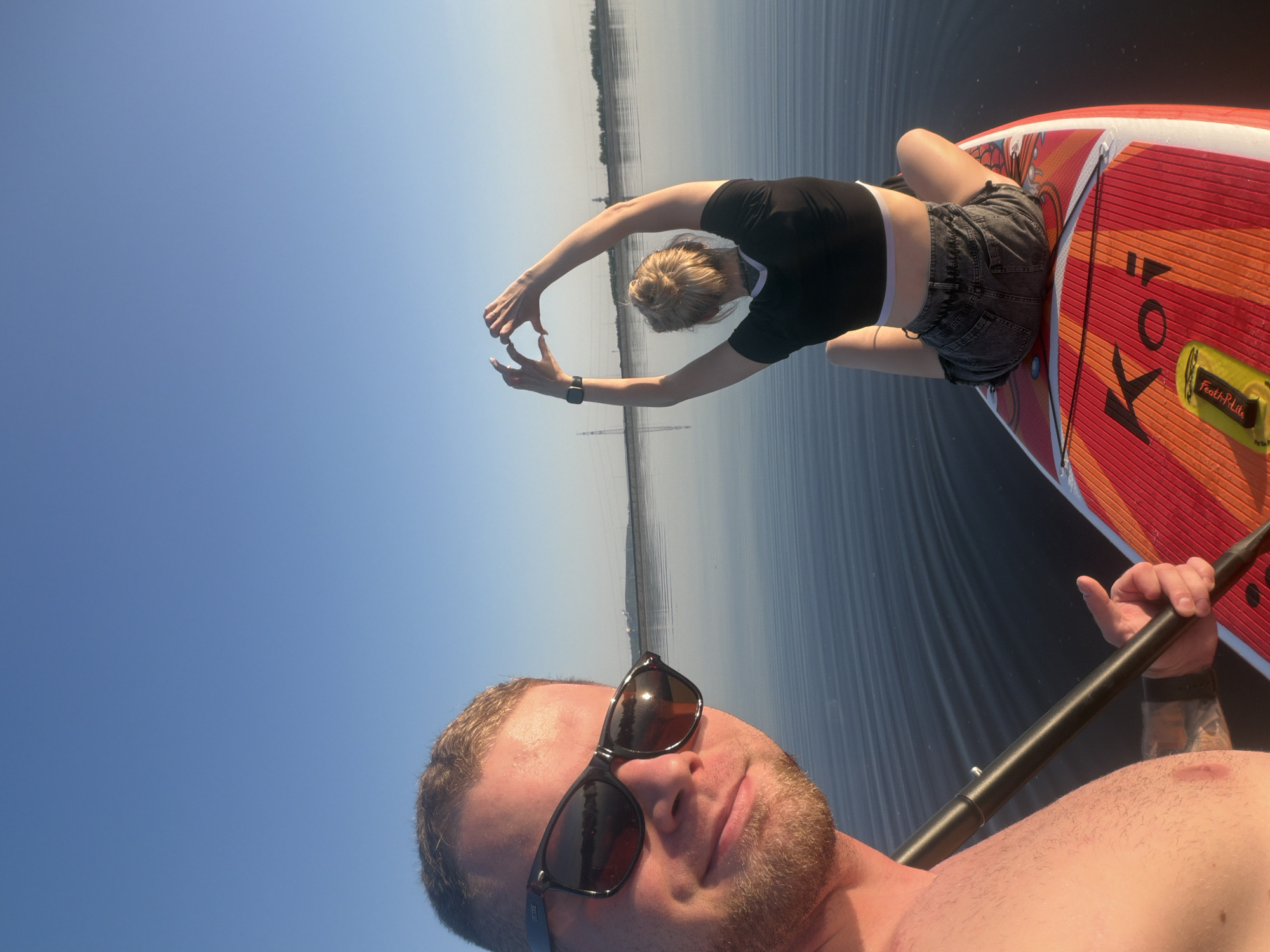 A low-angle, horizontally-oriented medium shot shows two people on what appears to be a stand-up paddleboard (SUP) on a body of water. The person in the foreground, likely holding the camera, is a shirtless man with light skin and short, light-brown hair. He is wearing dark sunglasses, and his face is angled slightly towards the viewer. He is holding a paddle.
Behind him, a second person, a young woman with her blonde hair in a bun, is kneeling on the paddleboard, facing away from the camera. She is wearing a black, short-sleeved crop top and grey shorts. Her arms are raised overhead, with her hands almost touching, forming an arch. The paddleboard is red and orange, with the words "KOI" visible on it.
The water is calm and reflects the sky, which is a clear, bright blue. In the distance, a treeline is visible. Transmission towers rise up on the horizon. Based on the environment, it is highly likely that this image was taken in Chaikovsky, Russia. The scene suggests a recreational activity on a calm, sunny day.