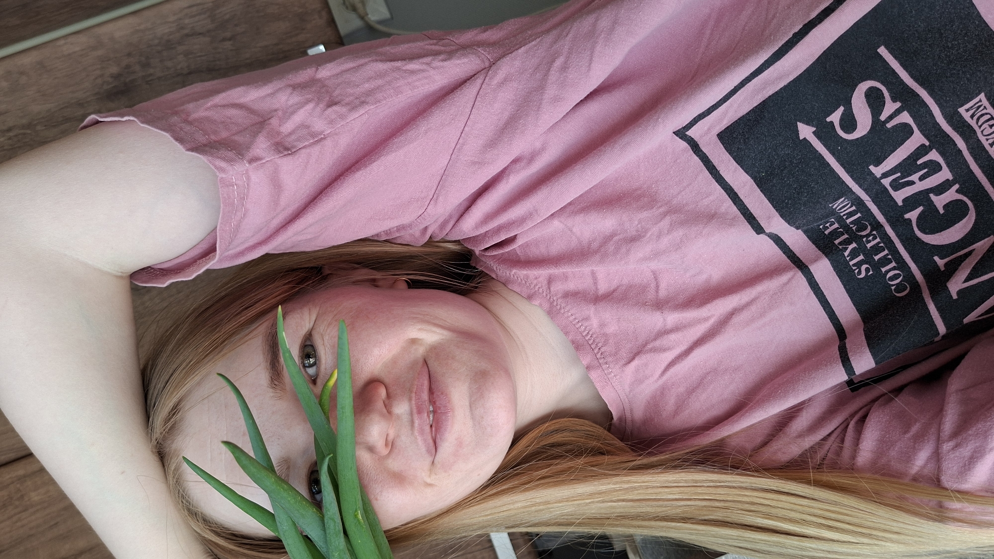 A woman lies on a wooden surface, possibly a floor. She wears a pink t-shirt with a black graphic reading "ANGELS STYLE COLLECTION GDM." Her blonde hair is spread out on the surface. She holds a bunch of green onions close to her face, partially obscuring it. The location is Большой Букор, Russia.