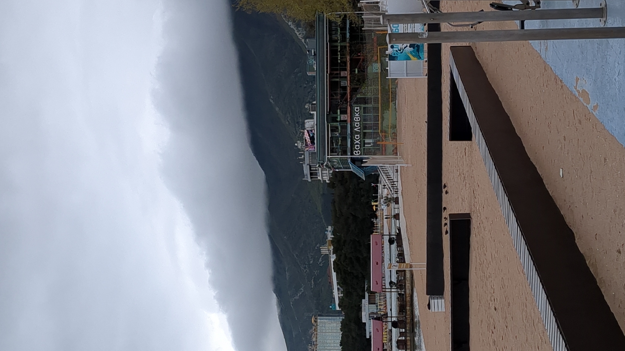 The image depicts a view in Gelendzhik, Russia. A mountain covered in low clouds is visible in the background. A multi-story building with the name "Βαχα Λαθκα" (likely a transliteration of a Greek name) is situated on the coastline. Adjacent to the building is a boardwalk or walkway near a beach. In the foreground, there is a low, tan-colored wall or structure with a dark brown or black trim running along its top edge, with periodic rectangular openings. Utility poles and other structures are also visible. The weather appears overcast.