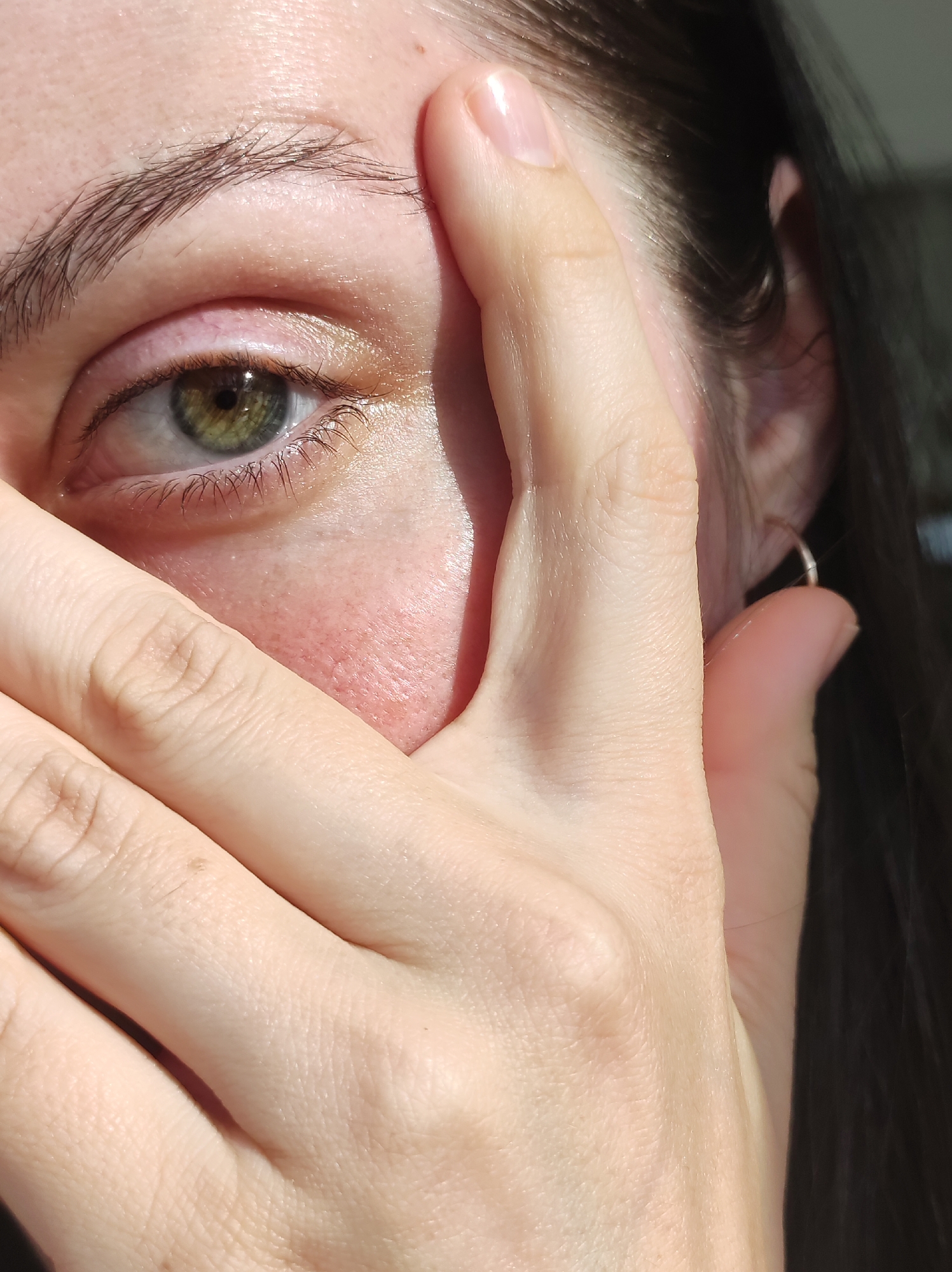 A close-up image shows the left side of a person's face, obscured by their own hand. The visible eye is green with a dark limbal ring. The eyebrow is dark and slightly arched. The skin tone is fair. The hand partially covers the face, with the fingers reaching towards the mouth. The fingernails are unpainted. A small silver hoop earring is visible in the earlobe. The hair is dark. The location is not identifiable from the image.