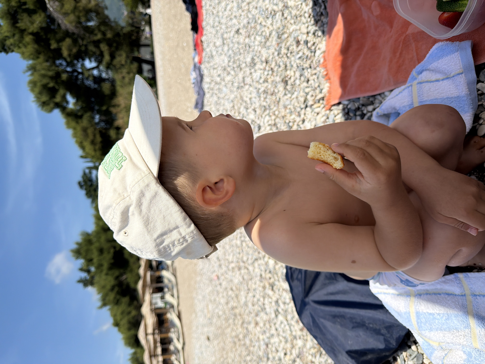 A toddler is sitting on a pebbled beach in Bečići, Montenegro, wearing a light-colored baseball cap. The child is holding a piece of bread in one hand, and is sitting on a blue and white striped towel, which is partially covering a dark blue sheet or blanket. Beside the towel is an orange towel and a plastic container with food visible inside. The backdrop is a beach with light-colored pebbles, foliage, buildings, and a blue sky.