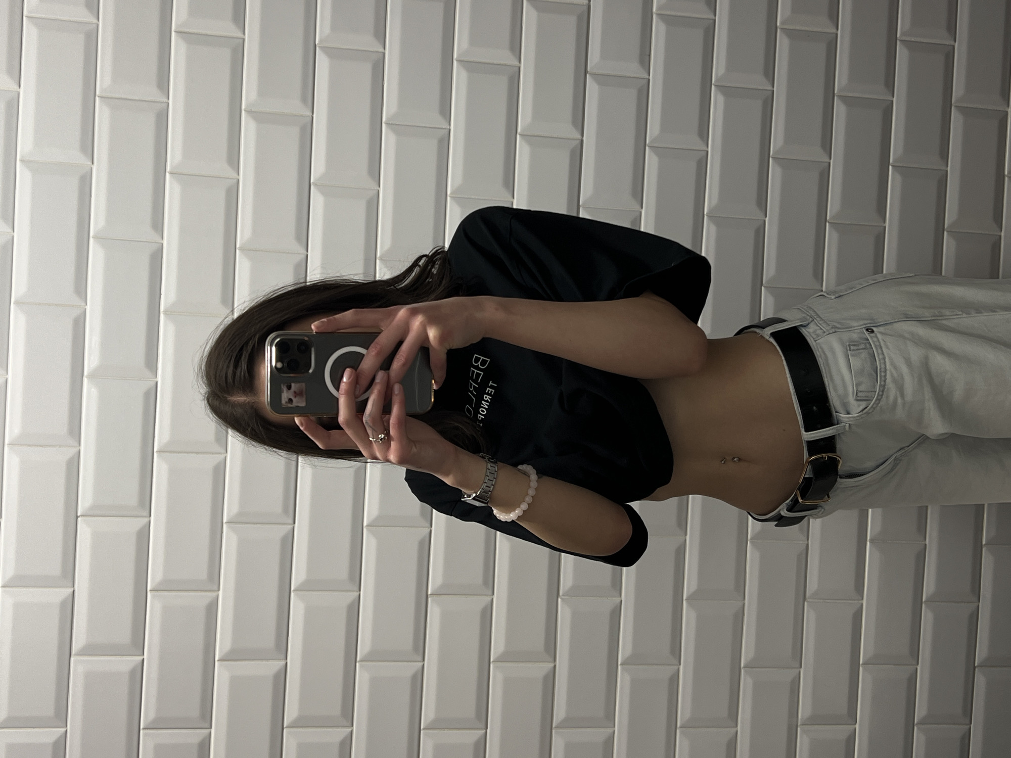 A young woman is taking a mirror selfie in a bathroom with white subway tiles. She is wearing a black crop top, light-wash jeans, and a black belt. She has a watch on her left wrist and a beaded bracelet on her right wrist. A silver ring is visible on her right index finger. The woman's belly button piercing is visible. The phone she is using to take the selfie has a phone case with a cat sticker. The reflection in the mirror shows the text "BEHIND" upside down on her shirt. The scene is likely in a private residence or public restroom in Ternopil, Ukraine, given the available context.