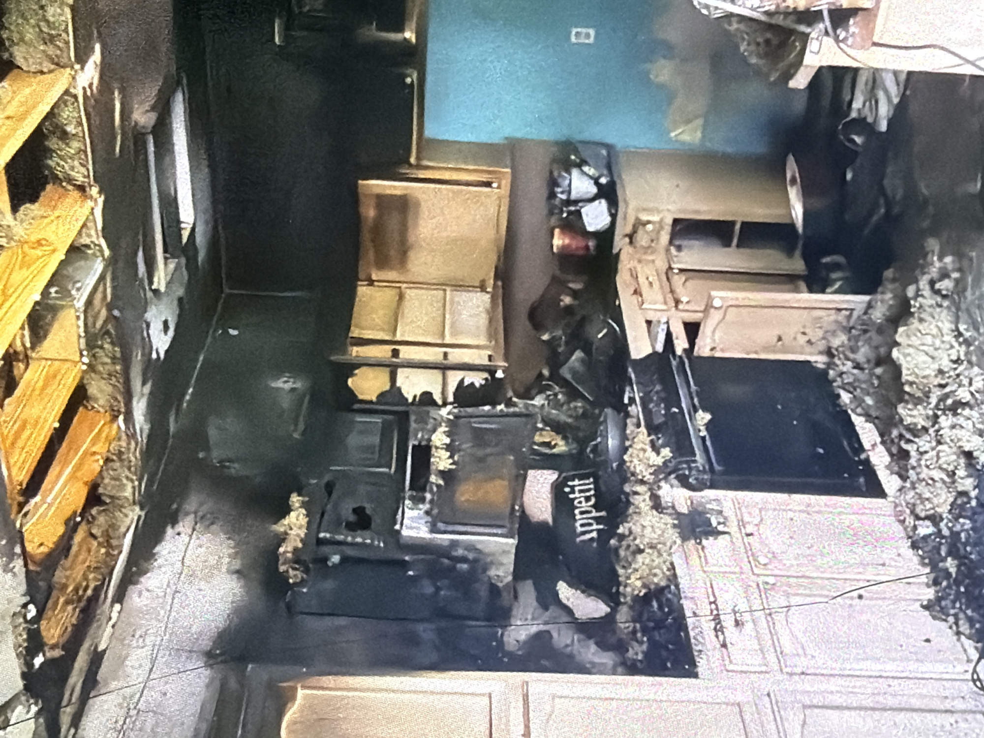 The image depicts the aftermath of a fire in a kitchen, likely in Bukuru, Nigeria. The scene is characterized by extensive fire damage. The ceiling is partially collapsed, revealing wooden beams and insulation. The walls and cabinets are heavily charred and blackened. A stove and oven appear damaged, with debris scattered around them, including what looks like insulation material and ash. A black fabric item with the word "appetit" is visible on the counter in front of the stove. There are open cabinets above the stove, some of which appear discolored. To the right, a teal-colored appliance, possibly a refrigerator, stands against a wall with an electrical outlet visible. The floor is covered in debris, ash, and what appear to be partially burned items. The overall impression is one of severe destruction within a domestic kitchen environment.