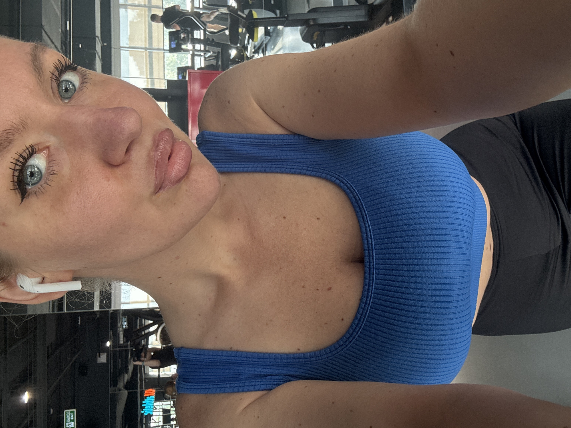 A low-angle, close-up selfie shows a young woman with light skin, light eyes, and blonde hair, wearing a blue ribbed sports bra and black leggings. An earbud is visible in her left ear. The background indicates a gym setting with exercise equipment and other people. The location appears to be indoors; Moscow, Russia is not visually confirmed.