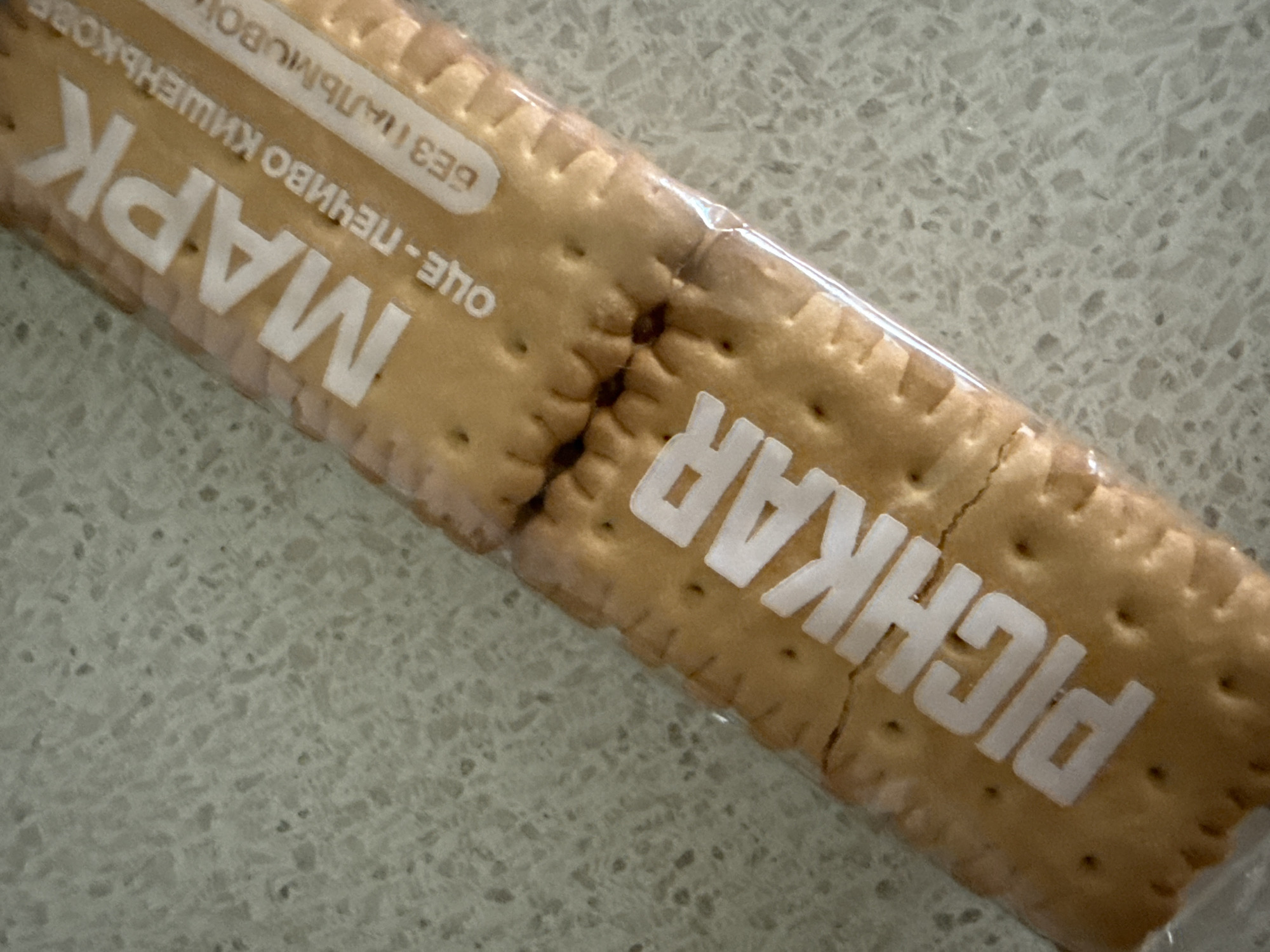 A package of biscuits is shown on a speckled, light-colored countertop. The package is clear plastic, revealing two rows of biscuits. White text is printed on the package in a diagonal orientation. The text on the upper half of the package reads "MAPK OUE-ПЕЧИВО КИШЕНЬКОВ БЕЗ ПАЛЬМОВОЇ". The text on the lower half of the package reads "РICHKAR". The biscuits themselves are rectangular with crimped edges and small indentations.
