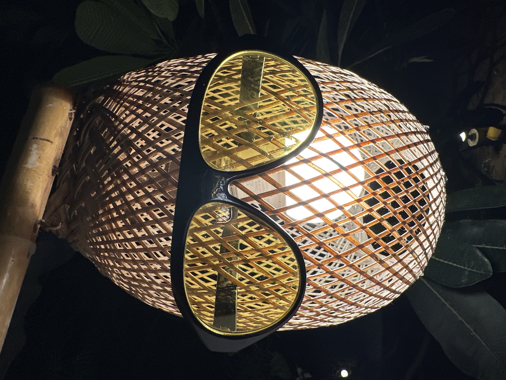 Captured in ตำบลเกาะพะงัน, Thailand, the image features a woven lamp shade affixed to a bamboo pole. The spherical lamp shade is made from a light brown woven material arranged in a grid pattern. The illumination within casts a bright glow through the latticework. Affixed to the shade are what appear to be over-sized sunglasses. The frames are dark, and the lenses have a golden hue. The scene is set at night, with surrounding greenery providing a backdrop.