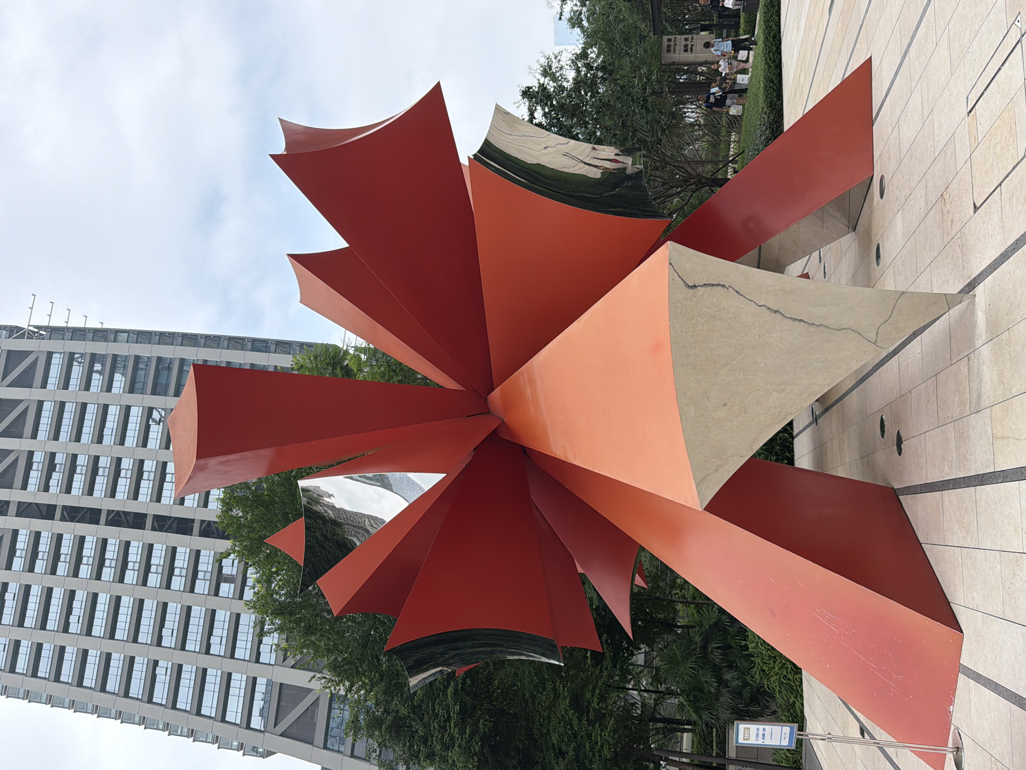 In Yuzhong District, China, a large, red, abstract sculpture with mirrored elements stands prominently in an outdoor plaza. The sculpture features sharp, angular planes that radiate outwards from a central point, with some sections appearing to curve inwards. The mirrored surfaces reflect the surrounding environment, including trees and a tall, modern building with a grid-like facade. The plaza pavement is tiled in a light color with dark linear inlays. Several trees are visible behind the sculpture, and a few people are present in the background, some standing near a sign with Chinese characters. The sky is overcast.