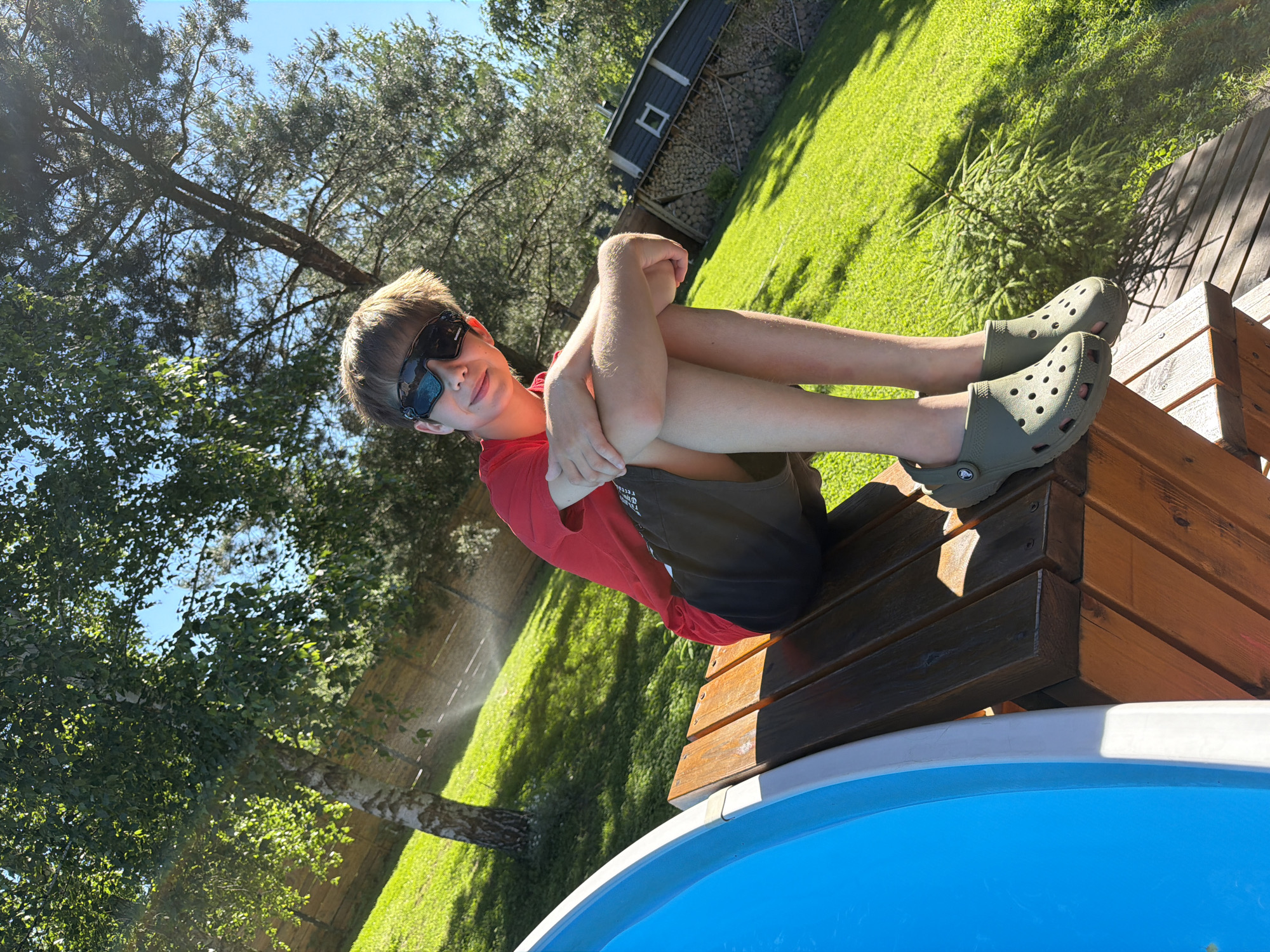 A young male, likely a pre-teen or early teenager, is seated on a wooden structure, possibly a bench or part of a deck, in Chertovitsy, Russia, on a bright, sunny day. He is wearing a red short-sleeved t-shirt, dark-colored shorts, and olive green Crocs. His head is slightly tilted, and he wears dark, wrap-around sunglasses. His forearms are crossed and resting on his bent knees, with his hands clasped or interlaced. He looks towards the camera with a subtle smile.
To the left of the wooden structure, a segment of a blue swimming pool with a white rim is visible. The background features a vibrant green, sloping lawn and various trees under a clear blue sky. A prominent birch tree with white bark stands on the left, while other conifer and deciduous trees are scattered throughout the scene. A wooden fence stretches horizontally in the mid-background. In the far right background, a dark-colored shed with a white-trimmed square window is visible, accompanied by a large, neatly stacked pile of firewood. Strong sunlight creates distinct shadows and a subtle lens flare in the upper left and lower right portions of the image. The entire scene is captured with a noticeable counter-clockwise tilt.