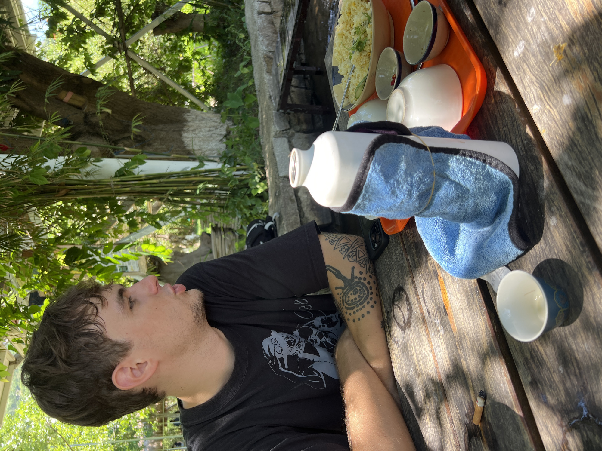 In an outdoor setting, likely a restaurant in Nam Cam Ranh, Vietnam, a young man with brown hair and tattoos on his left arm is seated at a wooden table, looking to his right. He is wearing a black t-shirt with a graphic print. In front of him on the table is a lit cigarette, a small blue and white cup, and a water bottle wrapped in a blue towel. Adjacent to the water bottle is an orange tray holding several white bowls, one of which contains a yellow, rice-based dish. The background shows lush green foliage, bamboo stalks, and trees, suggesting a tropical or subtropical environment. Sunlight filters through the leaves, casting dappled shadows on the table.