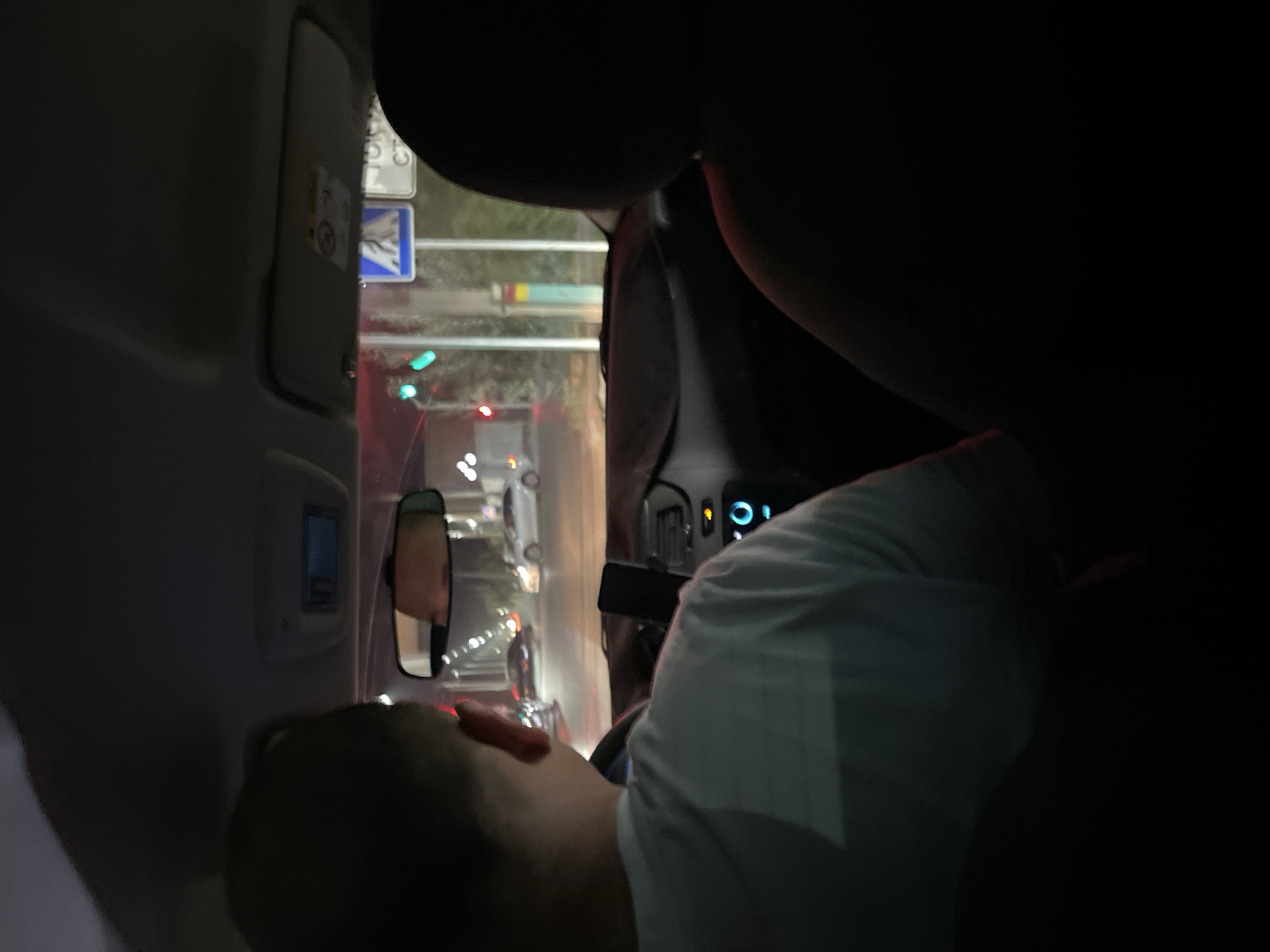 The image depicts the interior of a vehicle, viewed from the back seats, looking forward through the windshield. The scene appears to be at night in an urban environment, likely Kyzylorda, Kazakhstan, as indicated by the presence of traffic lights and road signs consistent with the region.
In the foreground, the back of a person's head and shoulders are visible on the left, facing forward and slightly to the right. This individual is wearing a light-colored t-shirt. On the right side of the frame, the silhouette of another person's head and shoulders is visible, also facing forward.
The dashboard of the car is visible, with a mobile phone mounted on a stand in the center. A circular illuminated control or button is present on the right side of the dashboard. The rearview mirror shows a reflection of a person's face.
Outside the vehicle, through the windshield, a road is visible with several cars. Traffic lights are illuminated, showing green and red signals. Road signs are present, including a blue sign with a pedestrian crossing symbol and another sign with Cyrillic text, which is partially obscured. The background suggests a city street with buildings and lights. The overall lighting conditions indicate it is nighttime.