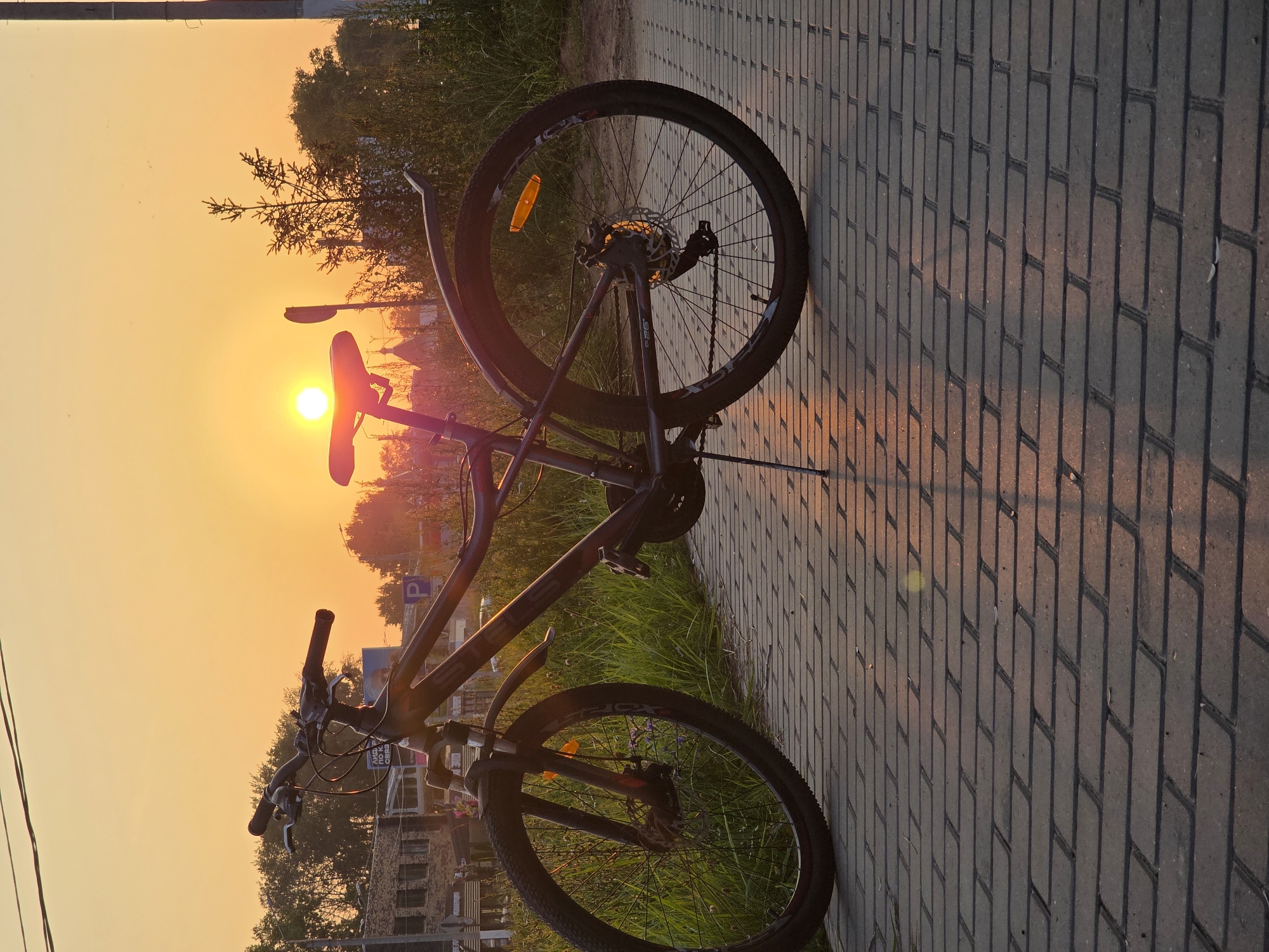 A dark-colored bicycle is parked on a paved walkway in front of a grassy area with trees in the background. The sun is setting, casting a warm orange glow across the sky and reflecting on the pavement. The bicycle is a mountain bike with a dark frame, black tires, and a red accent on the front fork. It has a kickstand supporting it. In the distance, there are buildings and what appears to be a church steeple. The lighting suggests it is late afternoon or early evening.