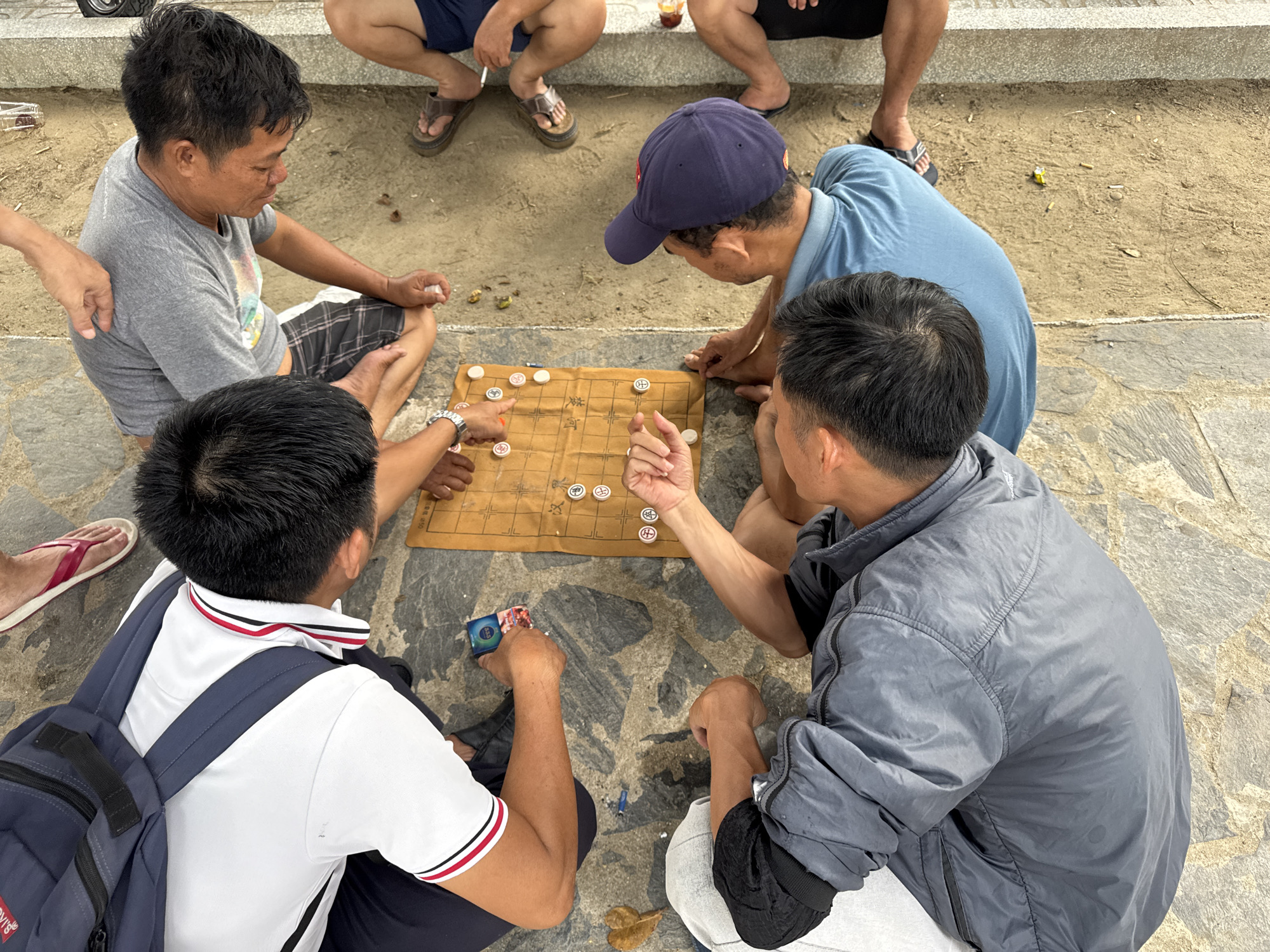 An overhead view captures a group of four men engaged in a game of Xiangqi, or Chinese chess, on a mat laid out on a cobblestone and sand surface, likely in a public area of Bac Nha Trang Ward, Vietnam. The players are seated around the board, their hands positioned to move the pieces. Three of the men are visible from the chest or waist up, while the fourth is mostly obscured. One man, wearing a grey t-shirt and plaid shorts, is on the left, his right hand holding a white chess piece. To his right, a man in a blue t-shirt and a navy baseball cap leans over the board, his left hand poised to make a move. In front of the man in the blue shirt, another man wearing a white polo shirt with red and black trim and a dark backpack is seated, his left hand holding a blue package. The fourth player, dressed in a grey jacket, is on the right, his body mostly visible as he participates in the game. The Xiangqi board is a traditional brown mat with a grid of lines and river. Scattered around the playing area are a few small objects, possibly debris or discarded items. The background shows a mixture of sand and grey stone, suggesting an outdoor setting, possibly a park or a promenade near a beach given the sandy texture. Several pairs of legs and feet are visible in the background, indicating other individuals are present but not directly participating in the game.