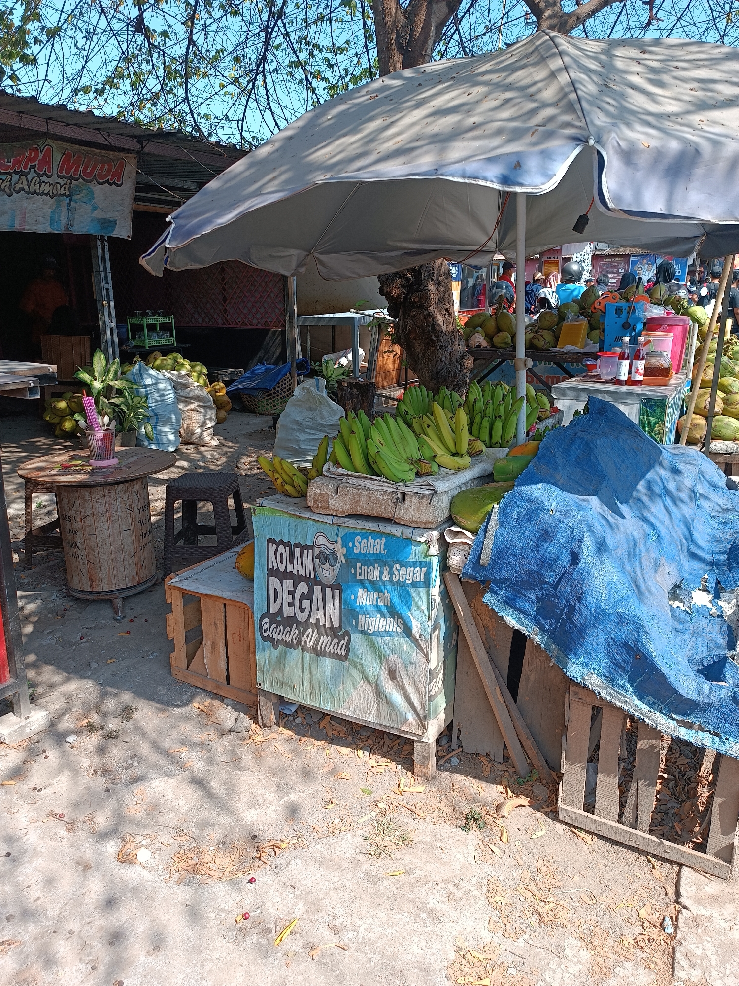 This image depicts an outdoor food stall in Imogiri, Indonesia, likely situated on a street or market area, as evidenced by the sunlit, dusty ground and the presence of various vendors and customers in the background. The stall is primarily focused on selling coconuts and bananas, with a prominent display of green bananas in bunches and numerous whole coconuts stacked and bagged. A large, light-colored umbrella provides shade over the stall.
In the foreground, a wooden table with a rustic design holds a pink cup with straws and a small plant. Adjacent to it is a small, dark plastic stool. A colorful banner with Indonesian text, including "KOLAM DEGAN" and "Bapak Ahmad," is displayed on a wooden structure. The banner also features text that appears to list product qualities like "Sehat" (Healthy), "Enak & Segar" (Delicious & Fresh), "Murah" (Cheap), and "Higienis" (Hygienic).
Behind the fruit display, several large, woven sacks filled with coconuts are visible. Other items on the stall include bottles of sauces or drinks, a pink jug, and a glass jar containing a brown liquid, possibly a beverage. In the middle ground, more coconuts are piled high, and a glimpse of other food stalls and customers can be seen under umbrellas. The overall environment suggests a busy, open-air market scene.