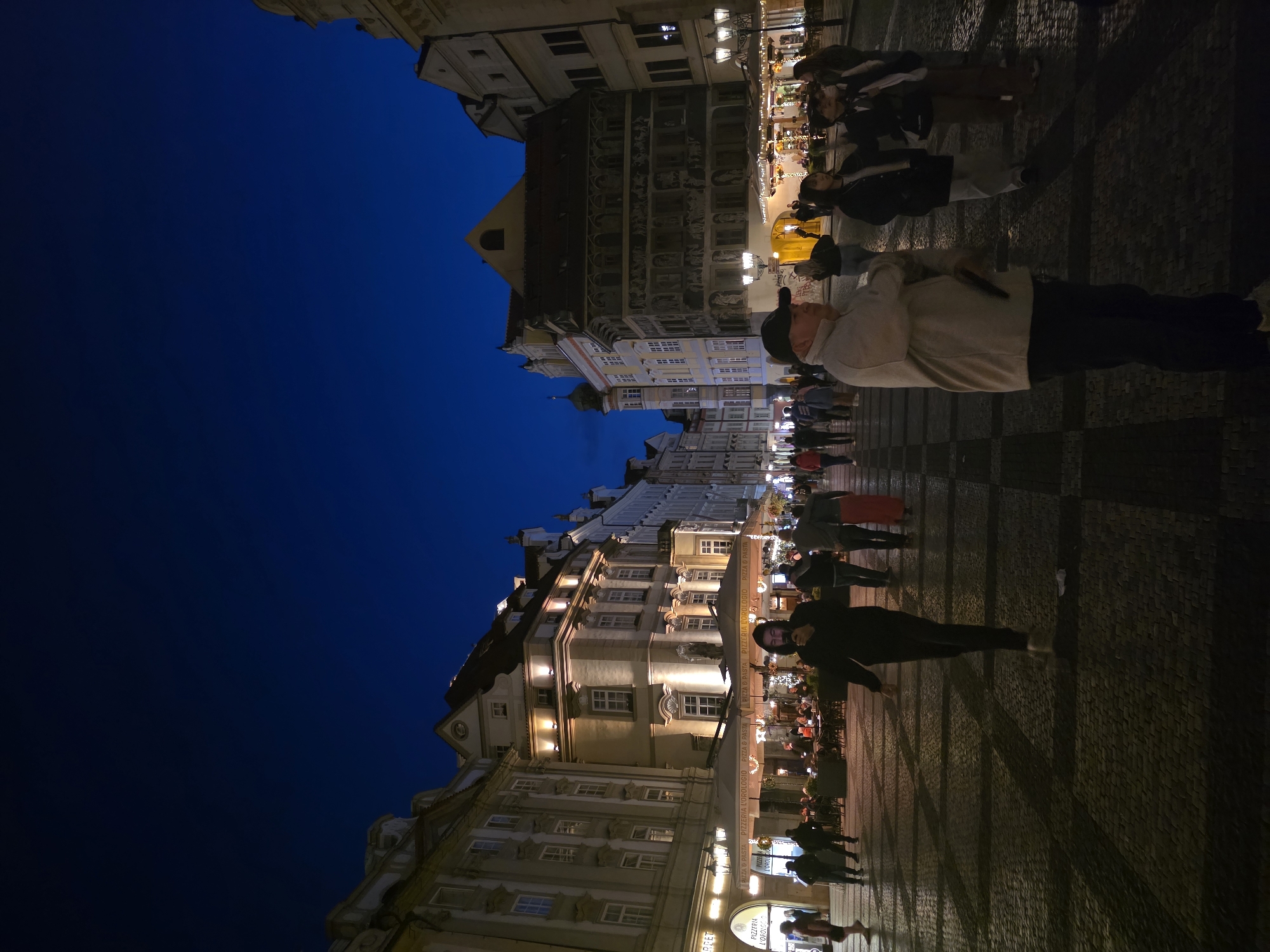 The image shows a street scene at night in an urban setting. The sky is a deep blue, indicating dusk or night. On either side of the street are tall, historic buildings with illuminated windows and architectural details. The street itself is paved with cobblestones and a pattern of darker rectangular stones.
Several people are walking or standing on the street. In the foreground, a man wearing a baseball cap and a light-colored jacket is facing away from the camera, with his right arm bent and holding something. To his left and further back, a person in a dark hooded jacket is walking towards the camera, looking down at their phone. Other individuals are dispersed throughout the scene, some walking in groups, others alone.
There are outdoor seating areas with umbrellas set up in front of some of the buildings, suggesting restaurants or cafes. Streetlights are visible, casting pools of light on the pavement. The overall atmosphere is lively and illuminated, despite being at night. The architecture suggests a European city.