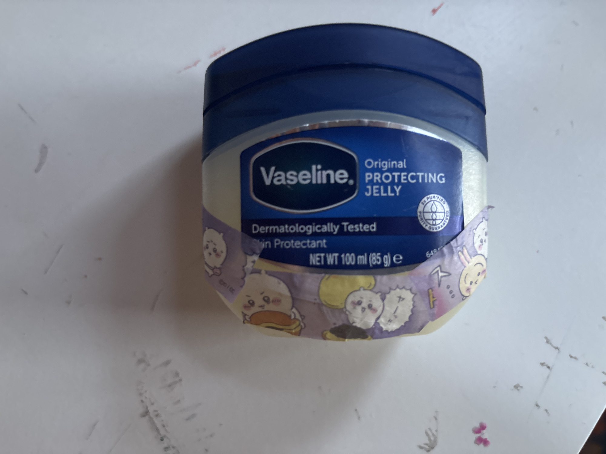 A Vaseline Petroleum Jelly container, decorated with a purple and white cartoon-themed tape, sits on a white surface. The label on the container is primarily blue and white. Text on the label reads "Vaseline. Original PROTECTING JELLY" and "Dermatologically Tested Skin Protectant NET WT 100 ml (85 g) e". The container is positioned at an angle, with the blue lid facing to the right and the label wrapping around the front. The white surface shows some smudges and faint pink marks.