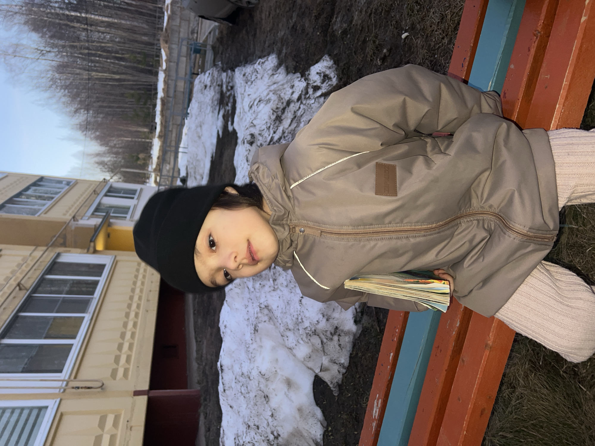 A young child wearing a black hat and a beige winter coat sits on a brightly painted bench outside a multi-story building. Patches of melting snow are on the ground. The location is likely Noviy, Russia, as indicated in the prompt. The child appears to be holding a stack of colorful books. Bare trees are visible in the background, suggesting late winter or early spring.