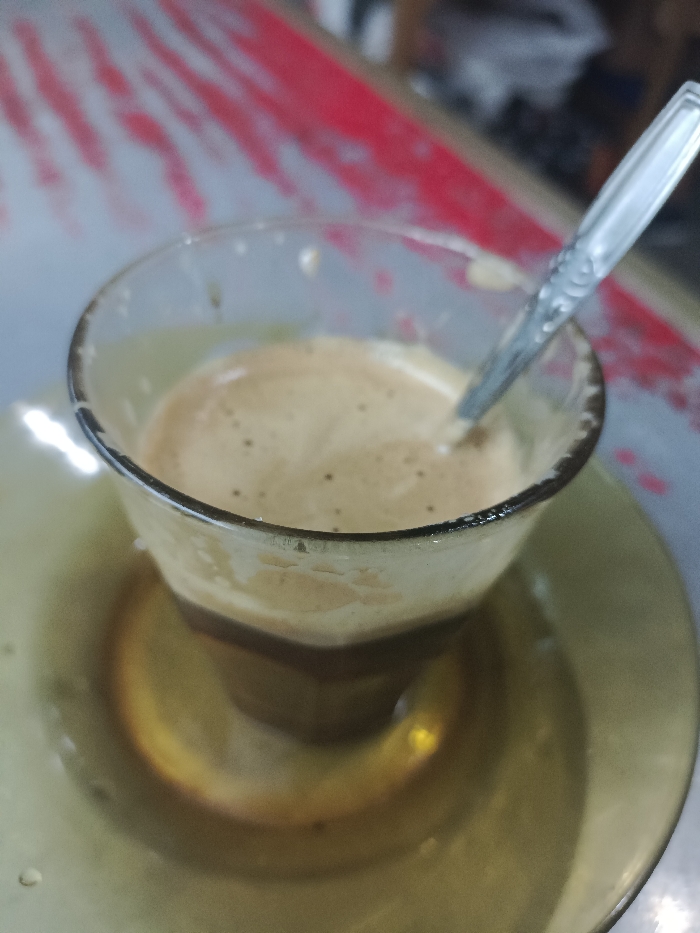 A glass of coffee with a foamy top sits on a greenish-yellow saucer. A metal spoon rests inside the glass, partially submerged in the coffee. The coffee appears layered, with a darker liquid at the bottom and a lighter, frothy layer on top. The background includes a blurred surface with red and white patterns. The overall composition suggests a close-up, casual shot of a beverage.