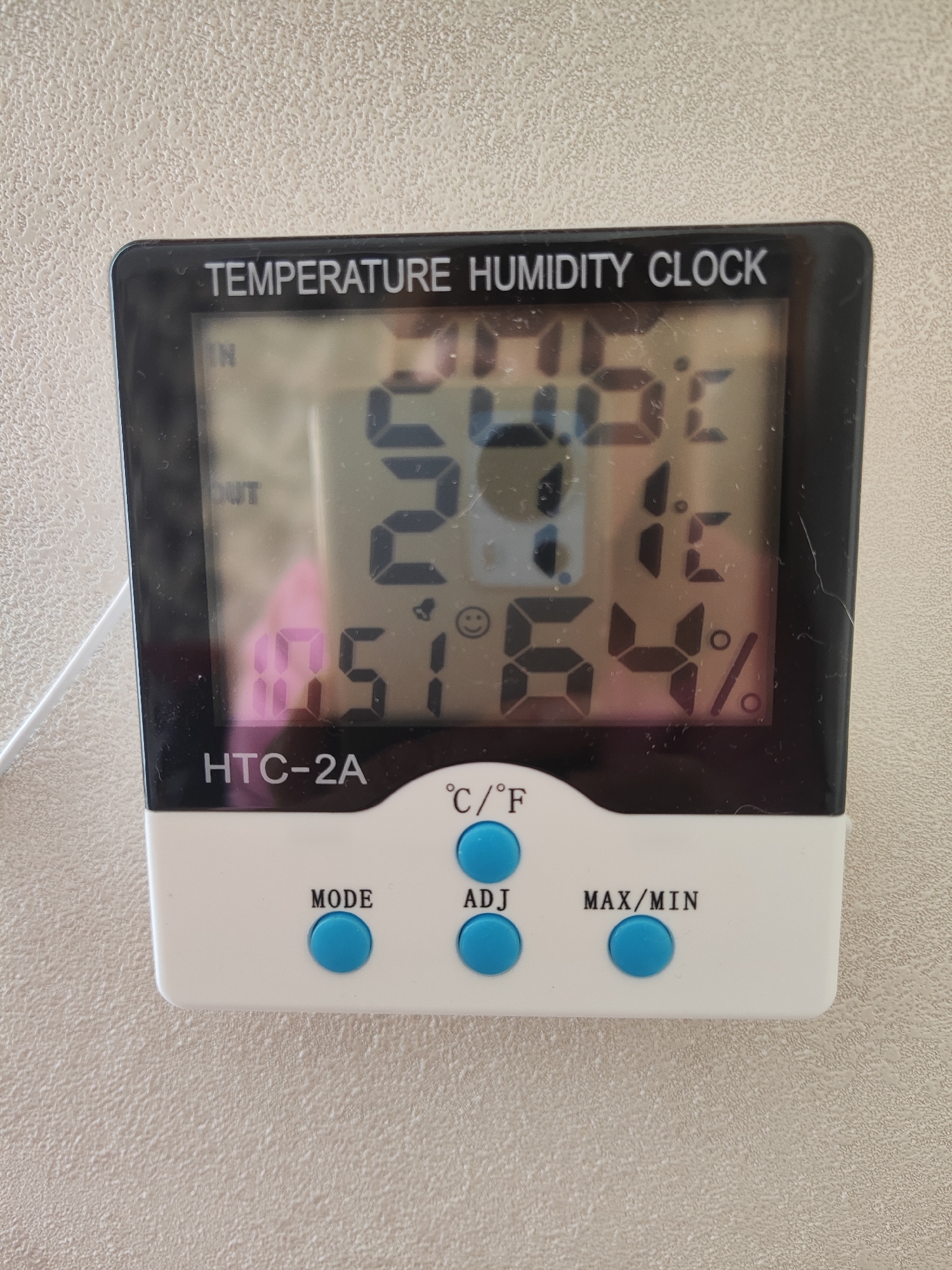 A close-up, eye-level shot shows a digital thermometer and hygrometer labeled "TEMPERATURE HUMIDITY CLOCK HTC-2A." The device is white with a black screen displaying readings. "IN" and "OUT" are visible on the screen, along with numerical values. The temperature displays as 70.8 degrees Fahrenheit, and the humidity reads 64%. Below the screen are three blue buttons labeled "MODE," "ADJ," and "MAX/MIN." The device is mounted on a textured, light-colored wall. The background is a neutral, lightly textured surface consistent with an indoor wall. The lighting is even, with no harsh shadows, suggesting an indoor setting. There are no people or animals visible in the frame.