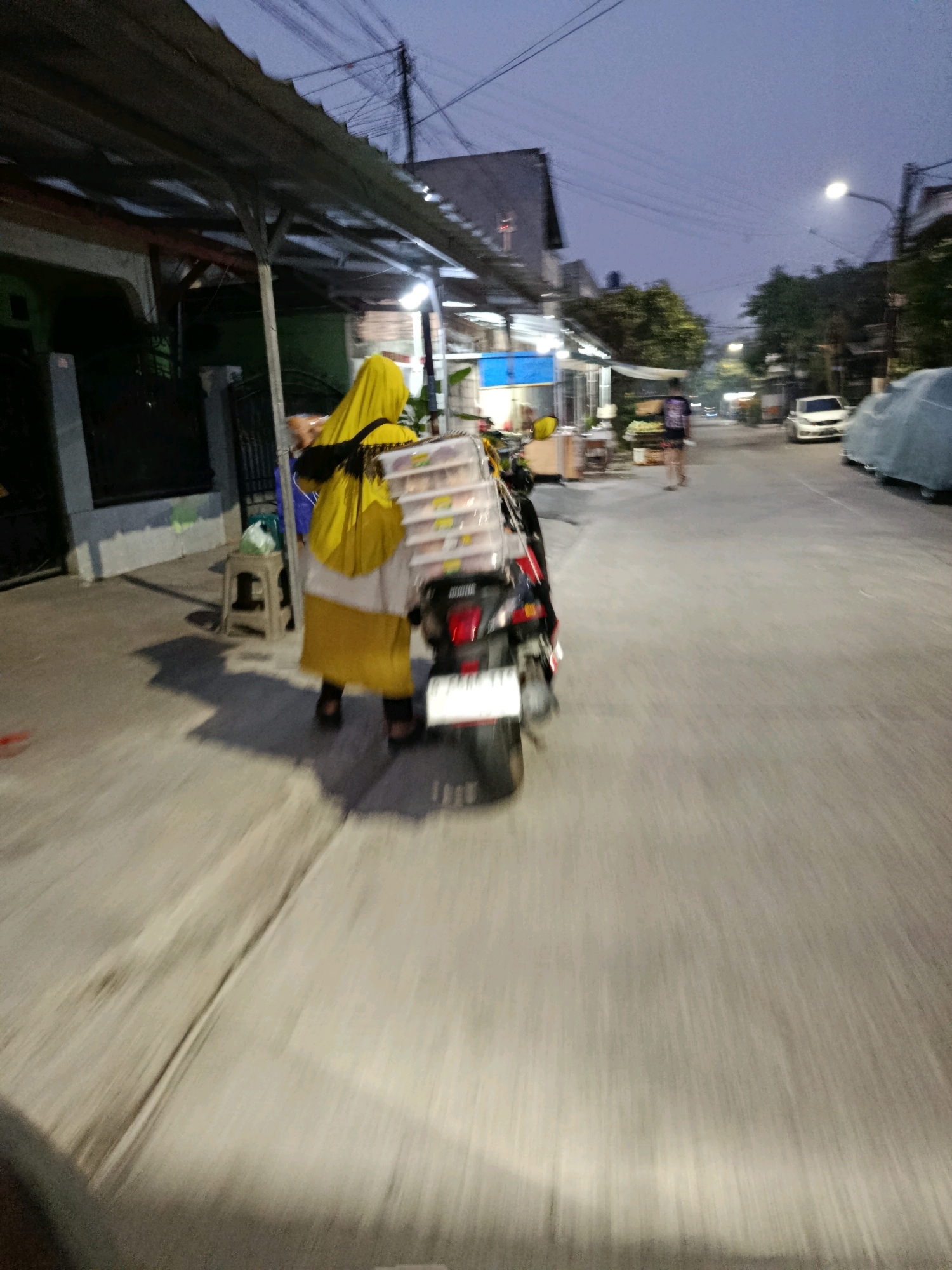 The image depicts a street scene in the city of Bekasi, Indonesia, during twilight. A person wearing a yellow hijab and a long yellow and white garment is riding a motorcycle loaded with stacked rectangular containers, likely food items. The person is moving away from the camera and is blurred due to motion. The motorcycle has a license plate that is also blurred. To the left of the person, there is a small stool and a green plastic bag. The street is paved and lined with buildings on both sides. Some of these buildings appear to be shops or food stalls with awnings and illuminated signs. There are trees and utility poles visible. In the background, other vehicles, including a white car and a covered vehicle, are present on the street. The sky is a dim, dusky blue. The overall impression is that of a residential or commercial street in a Southeast Asian urban environment.