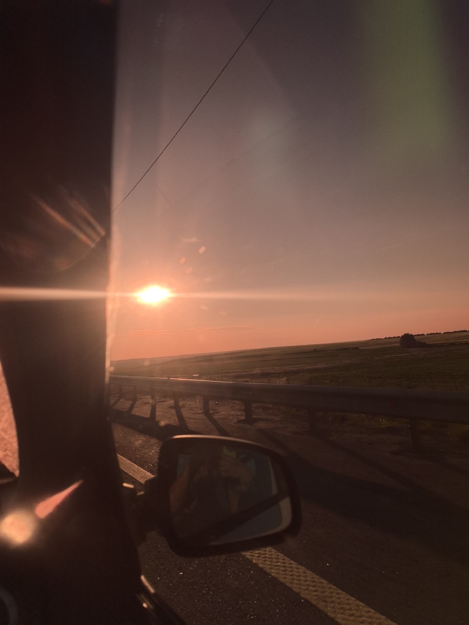 The image depicts a sunset viewed from inside a vehicle, likely on a highway or rural road. The sun, a bright orb with a lens flare, is positioned low on the horizon, casting a warm, orange glow across the sky and the landscape. The sky is a gradient of pink and orange, with a faint hint of green in the upper right corner, possibly from atmospheric scattering or a lens effect.
To the right of the sun, a vast, rolling landscape of green fields stretches to the horizon, dotted with a few scattered trees. A metal guardrail runs along the side of the road, separating the vehicle from the open fields. The road surface appears to be asphalt, with a yellow and white striped marking visible in the foreground.
In the foreground, the dark silhouette of the vehicle's A-pillar and side mirror dominates the left side of the frame. The side mirror reflects a partial view of the interior of the vehicle, showing a person's head and shoulders, possibly the driver or a passenger. The overall impression is one of a serene and picturesque journey during golden hour.
There is no indication of the city of Yumorga, Russia, or any specific geographical features that would place the image there.