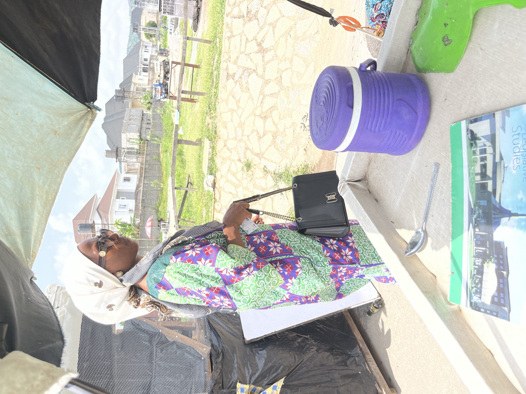 The image shows a middle-aged woman wearing a white headscarf, patterned dress, and sunglasses, sitting at an outdoor stall or table in what appears to be a market or roadside setting in Kubwa, Nigeria. The stall is covered by a black tarp. On the table next to her are a black purse with a chain strap, a purple cooler, a green plastic item, a book titled "Legislative Studies", and a spoon. The backdrop includes houses and a grassy area. The stall seems to be improvised, with black plastic sheeting underneath the table.