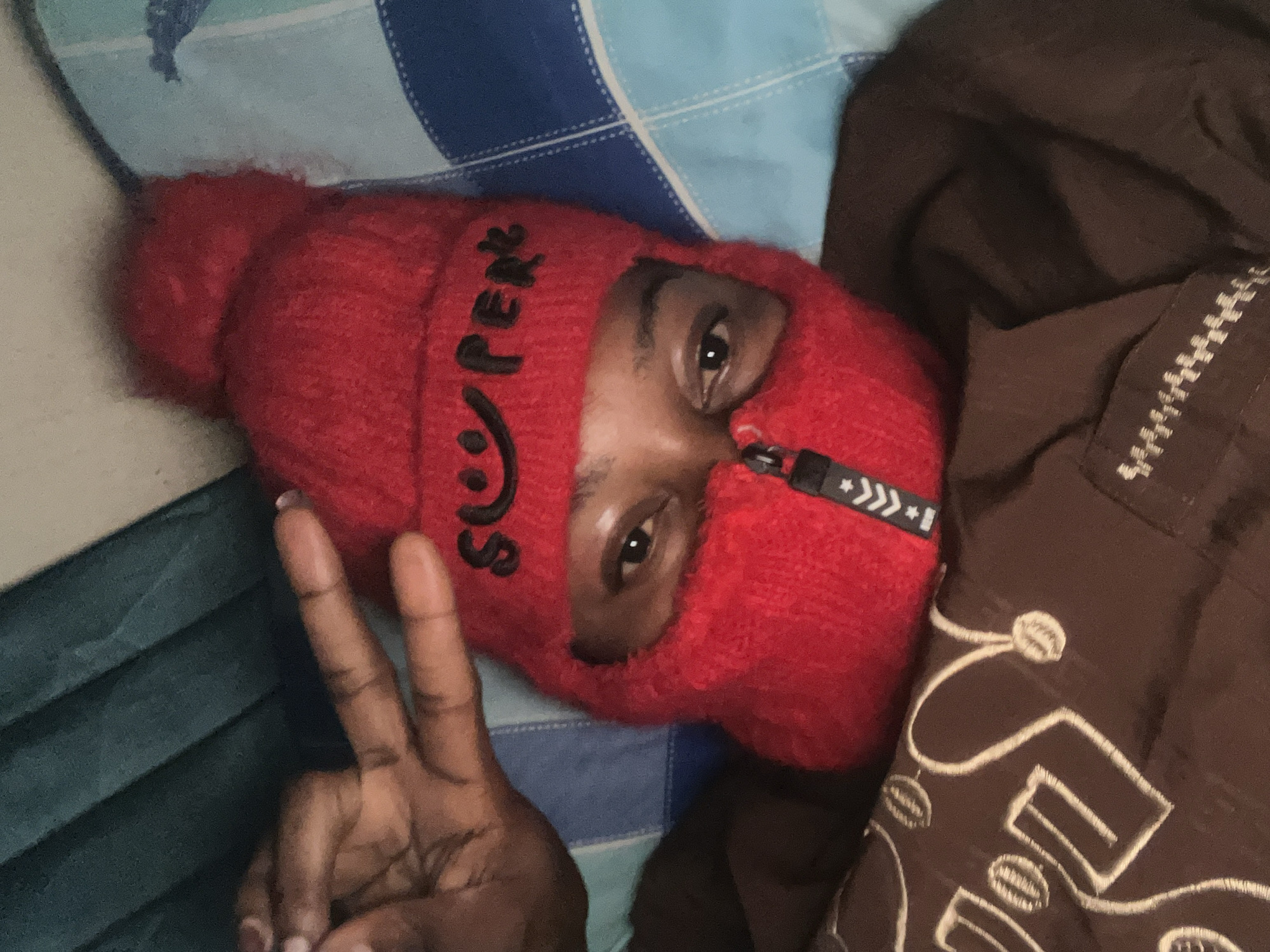 A close-up, overhead shot captures a person lying down, wearing a red knitted balaclava with a zipper and the word "SUPER" embroidered on it in black thread, along with a smiley face. The person's eyes are visible, and they are holding up two fingers in a peace sign with their left hand. The background consists of a blue and white striped surface, possibly a bed or pillow, and a dark green fabric. The individual is also wearing a brown garment underneath the balaclava, with white embroidery on the chest. The scene appears to be indoors, in a residential setting. There are no specific indicators to pinpoint the location to Jos, Nigeria.
