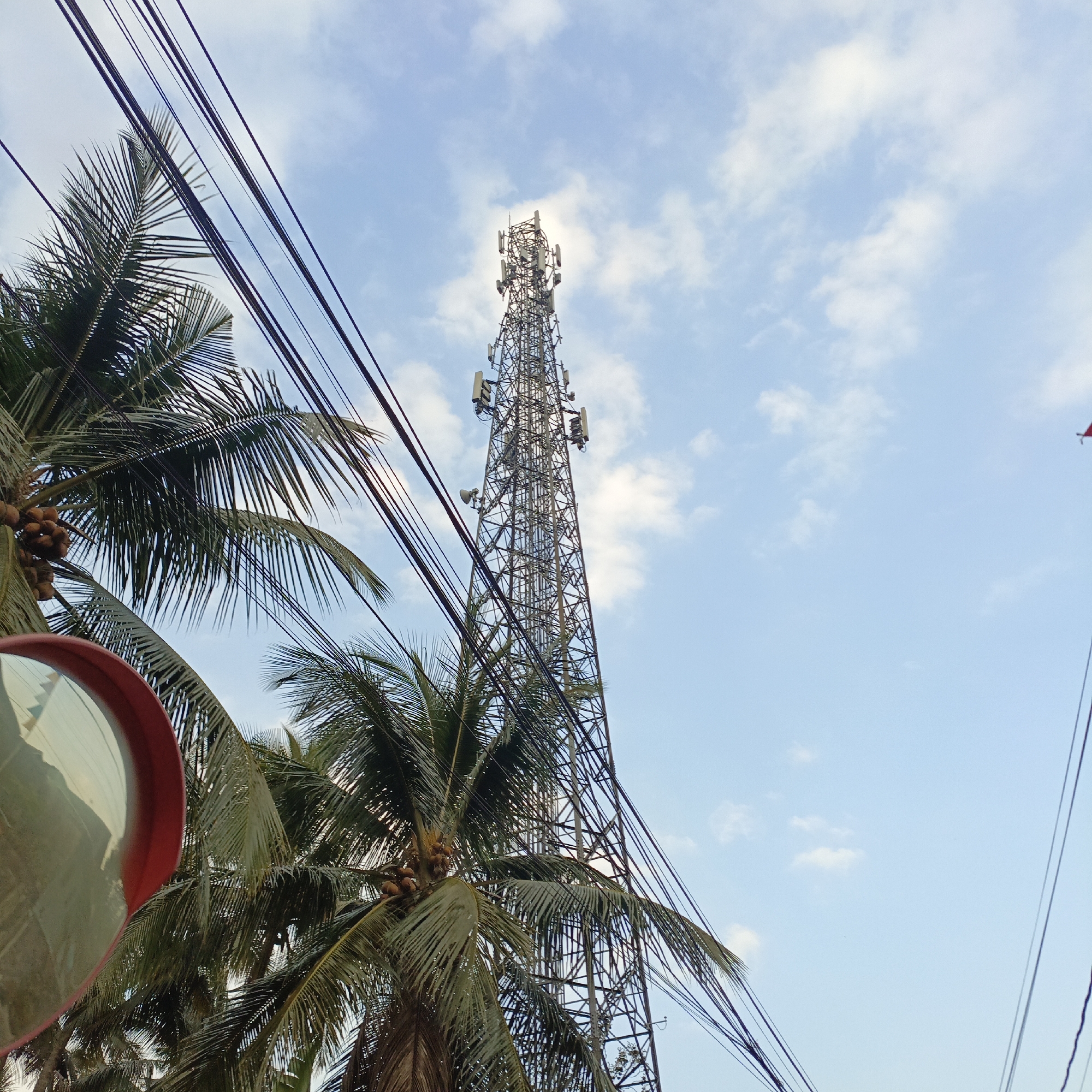 This is a low-angle, eye-level, outdoor, daylight, medium shot of a cell tower seen from a street level perspective in Salatiga, Indonesia. The cell tower is tall, made of steel lattice, and equipped with multiple antennas and dishes. It is positioned in the center of the frame, with its top obscured by the sun, creating a lens flare effect. In the foreground, on the left, a curved red-rimmed traffic mirror is visible, reflecting a distorted view of the surroundings, including palm trees and power lines. Several fronds of palm trees with coconuts are present on the left and center of the image. Numerous dark power or communication lines crisscross the frame, obscuring parts of the sky and the cell tower. The sky is a bright blue with scattered white clouds. The overall impression is one of urban infrastructure blending with tropical vegetation.