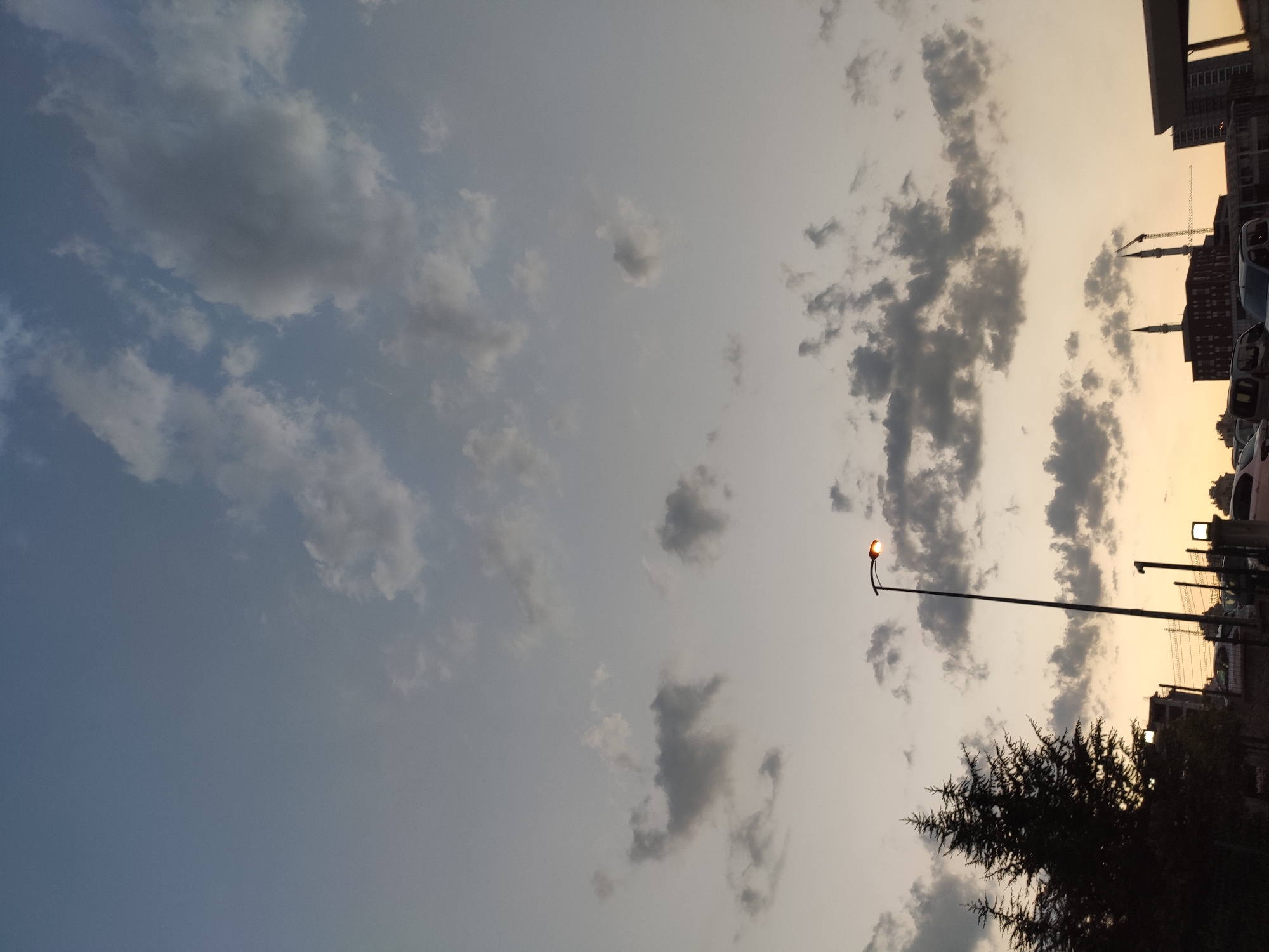 A streetlamp with an illuminated orange bulb stands to the right of center in this outdoor, low-angle shot of a sky at dusk. The sky is a gradient of pale blue at the top, transitioning to a muted yellow near the horizon. Scattered clouds, ranging in color from light gray to dark gray, drift across the sky. To the left, the dark silhouette of two evergreen trees is visible against the brighter sky. In the background, several buildings are partially visible, including one with two prominent spires resembling minarets, suggesting a mosque, and at least one construction crane. The lower portion of the image shows a street scene with parked cars and a fence. The overall lighting suggests a twilight or early evening setting. The image appears to have been taken in Etimesgut, Turkey.