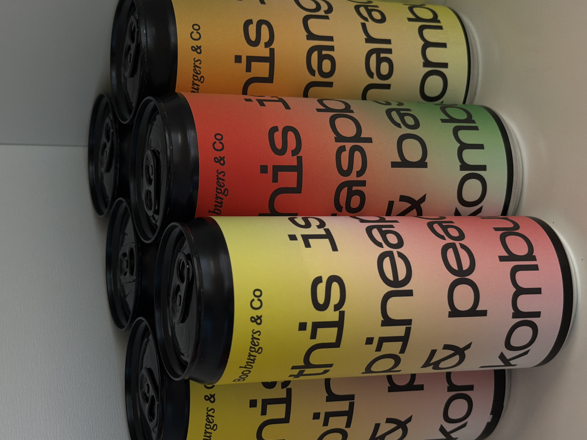 The image shows a stack of Booburgers & Co Kombucha cans. One flavor is mango & banana kombucha. Another is raspberry & baobab kombucha. The other is pineapple & peach kombucha. The cans are stacked in a white space. The location and city of Riga, Latvia, are not discernible from the image.