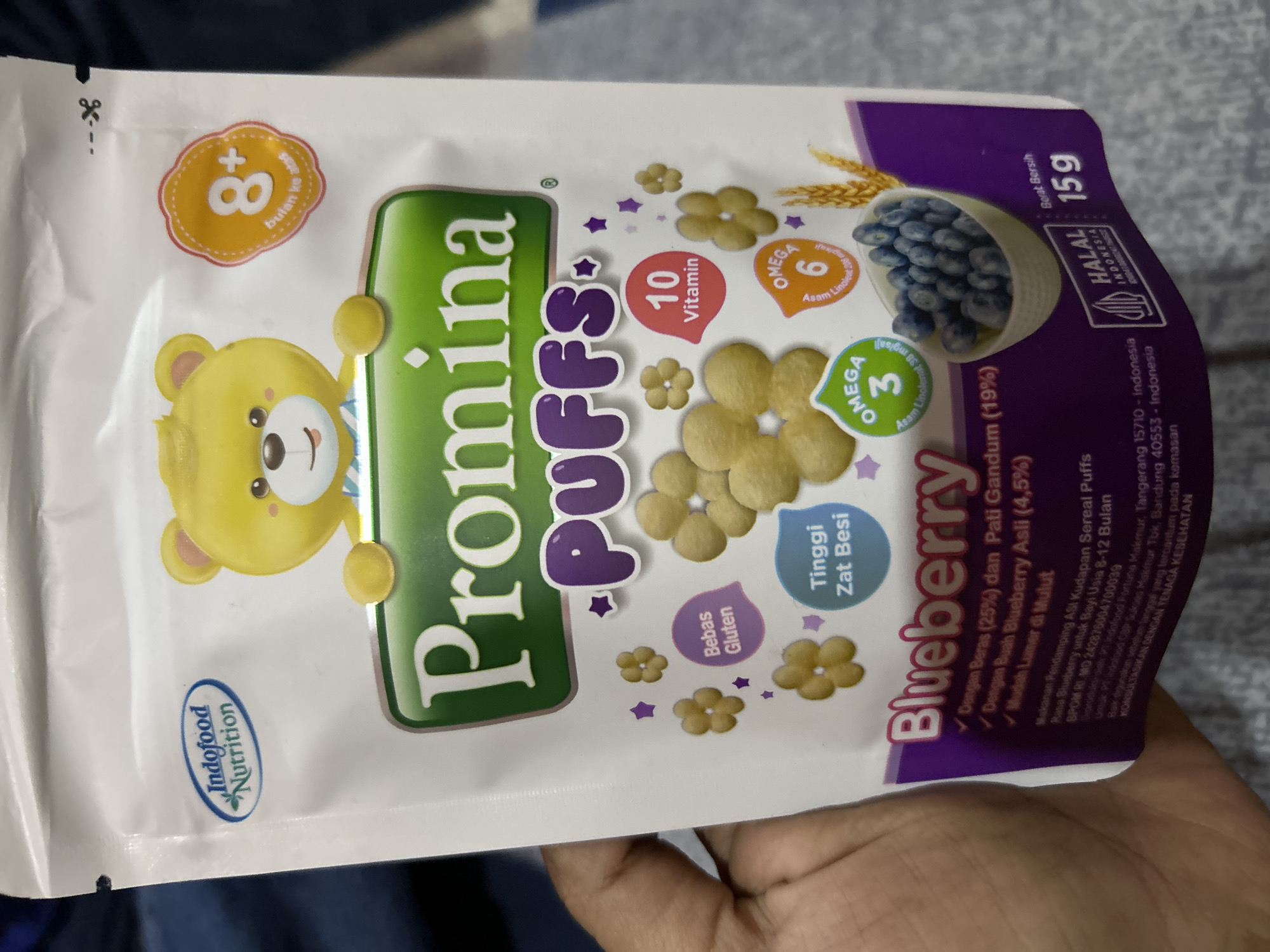 The image shows a close-up of a pouch of Promina Puffs Blueberry cereal for babies, held by a person's hand. The pouch is white with colorful branding. Prominently displayed are the brand name "Promina Puffs," a cartoon bear character, and text indicating "Blueberry," "8+ bulan ke atas" (8+ months and up), "Bebas Gluten" (Gluten-Free), "Tinggi Zat Besi" (High Iron), "10 Vitamin," "Omega 3," and "Omega 6." Nutritional information and company details are visible at the bottom of the pouch, including "PT. Indofood Fortuna Makmur, Tangerang 15110 - Indonesia" and "BPOM RI MD 242876004700039". The text "HALAL INDONESIA" is also present. The background is blurred and appears to be a surface, possibly a table or floor. There is no indication of the specific location within the city of Depok, Indonesia, other than the product itself which is manufactured in Indonesia.