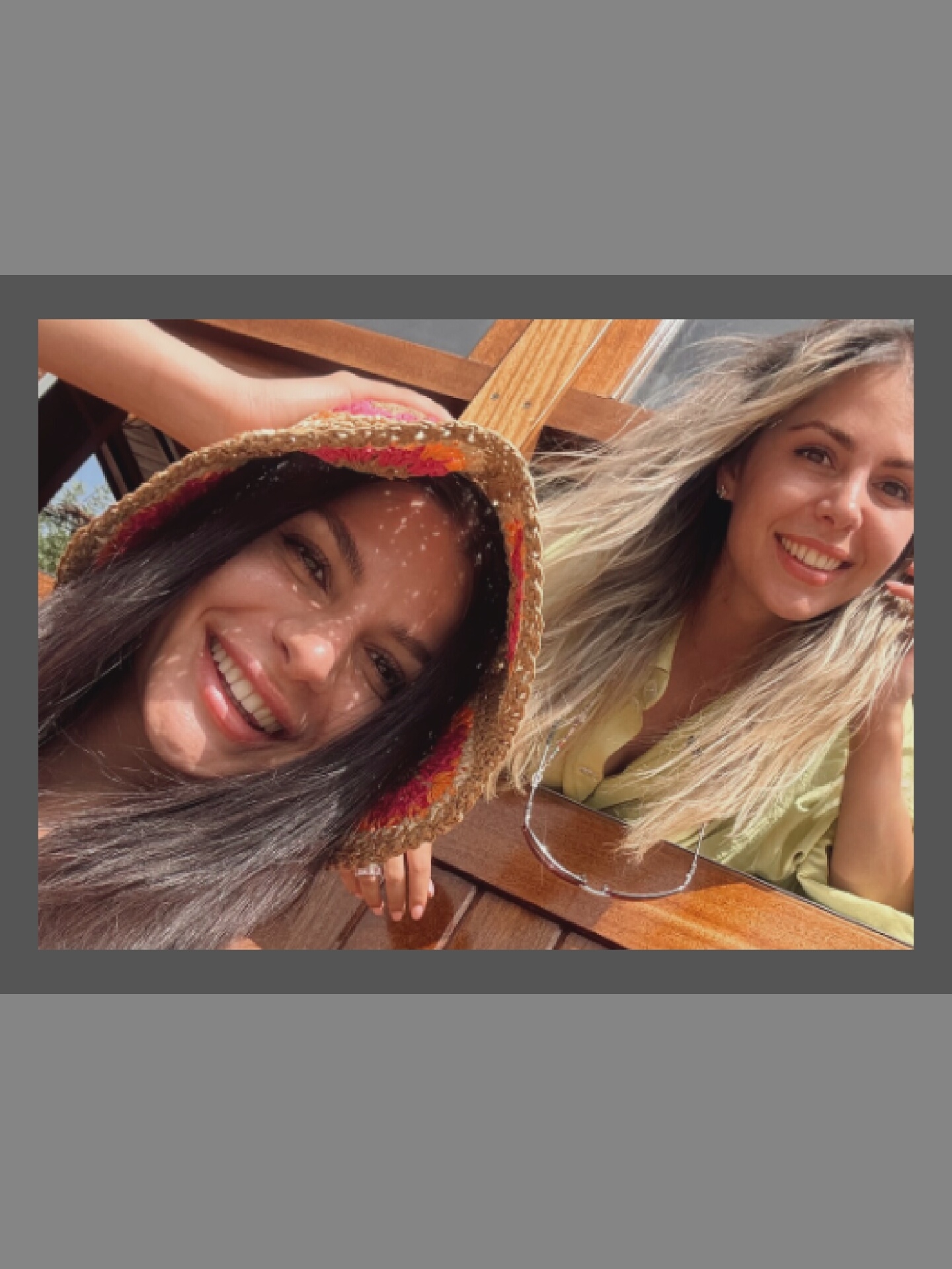 Two women are captured in a close-up selfie outdoors. The woman on the left is wearing a straw hat with colorful accents and is smiling broadly, with dappled sunlight falling on her face. Her long, dark hair frames her face. The woman on the right has long, blonde hair and is also smiling, wearing a light green top. Her eyes are brown, and she has small earrings. They appear to be sitting at a wooden table or bench. The background consists of wooden structures and possibly foliage, suggesting a casual outdoor setting.
