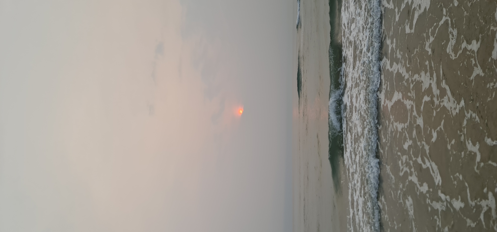 The image captures a tranquil coastal scene, likely a sunrise, with the camera oriented to present a horizontal landscape. The left side of the frame is dominated by a hazy, overcast sky, rendered in soft shades of grey and pale rose. Through a dense atmospheric haze and clouds, the sun appears as an intense, glowing orange-red disc, partially obscured but radiating a soft light.
Below the horizon, the ocean extends, reflecting the muted light. Gentle waves roll in, breaking into white foam as they approach the shore. The water appears a subdued grey-green. On the right side of the image, a stretch of wet, sandy beach is visible, intricately patterned by the receding white foam of the waves. No people or artificial structures are present, emphasizing the natural beauty of the moment.
The overall ambiance suggests a peaceful dawn in a coastal location like Đà Nẵng, Vietnam, where the sun rises over the sea. The weather is hazy or misty, diffusing the sunlight and creating a serene, almost ethereal atmosphere. There is no visible text in the media file.