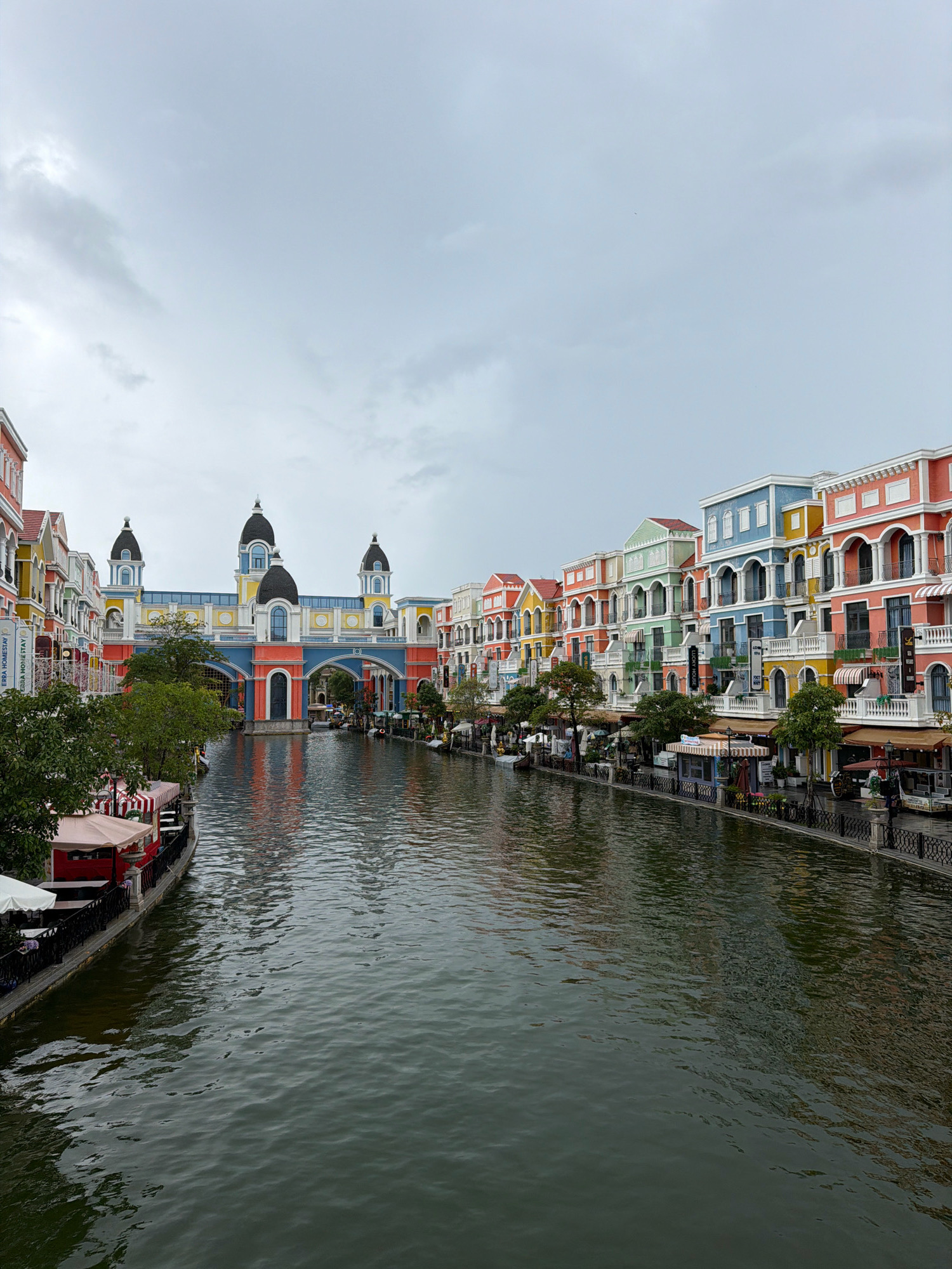 The image captures a picturesque canal lined with vibrantly colored buildings in Phu Quoc, Vietnam. The architecture is a mix of Italian Renaissance and French colonial styles, with a row of pastel-colored structures featuring balconies and decorative facades on the right bank. On the left bank, the buildings are similarly adorned, with awnings extending over pedestrian areas.
A prominent structure resembling a grand archway or bridge spans the canal in the mid-ground, connecting the two sides. This architectural piece is painted in bold hues of blue, yellow, and red, with domed towers crowning its upper sections.
The canal itself is the central element, with dark, rippling water reflecting the colorful buildings and the overcast sky. Several small boats are visible on the water, some moored along the banks and others potentially in motion. Pedestrians can be seen walking along the canal's edges, adding a sense of activity to the scene. Trees are interspersed along the walkways, providing pockets of greenery against the man-made structures.
The overall atmosphere is one of a charming European-inspired town, transplanted to a tropical setting, likely a tourist destination. The cloudy sky suggests overcast weather, but it doesn't detract from the visual appeal of the colorful architecture. This scene is characteristic of Sunset Town or Hon Thom Island in Phu Quoc, known for its Mediterranean-inspired architecture and waterfront attractions.
