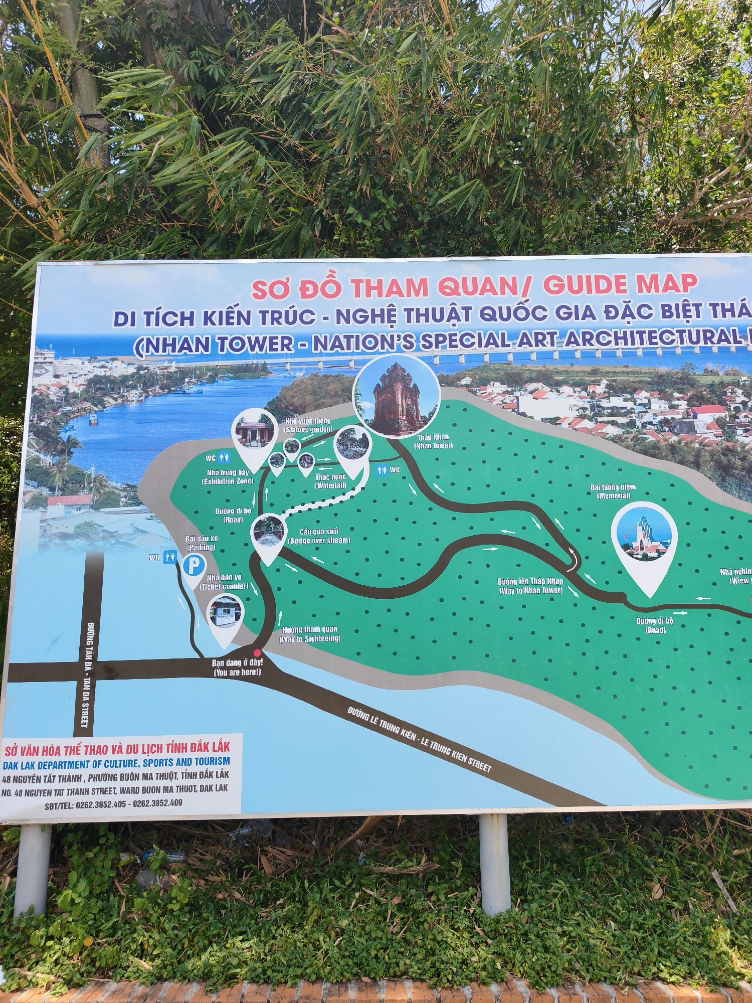 The image shows a large, rectangular sign displaying a map of the Nhan Tower historical and cultural site in Tuy Hoa city, Vietnam. The map is printed on a light blue and green background, with red and white text. It outlines various attractions within the site, including the Nhan Tower itself, a waterfall, a statues garden, a memorial, and a ticket counter. Pathways and roads are clearly marked. The sign also includes contact information for the Dak Lak Department of Culture, Sports and Tourism, suggesting the site might be related to that province, despite being located in Tuy Hoa. The sign is supported by two metal poles and is set against a backdrop of lush green vegetation and bamboo. The overall scene appears to be outdoors, likely in a public park or tourist area.