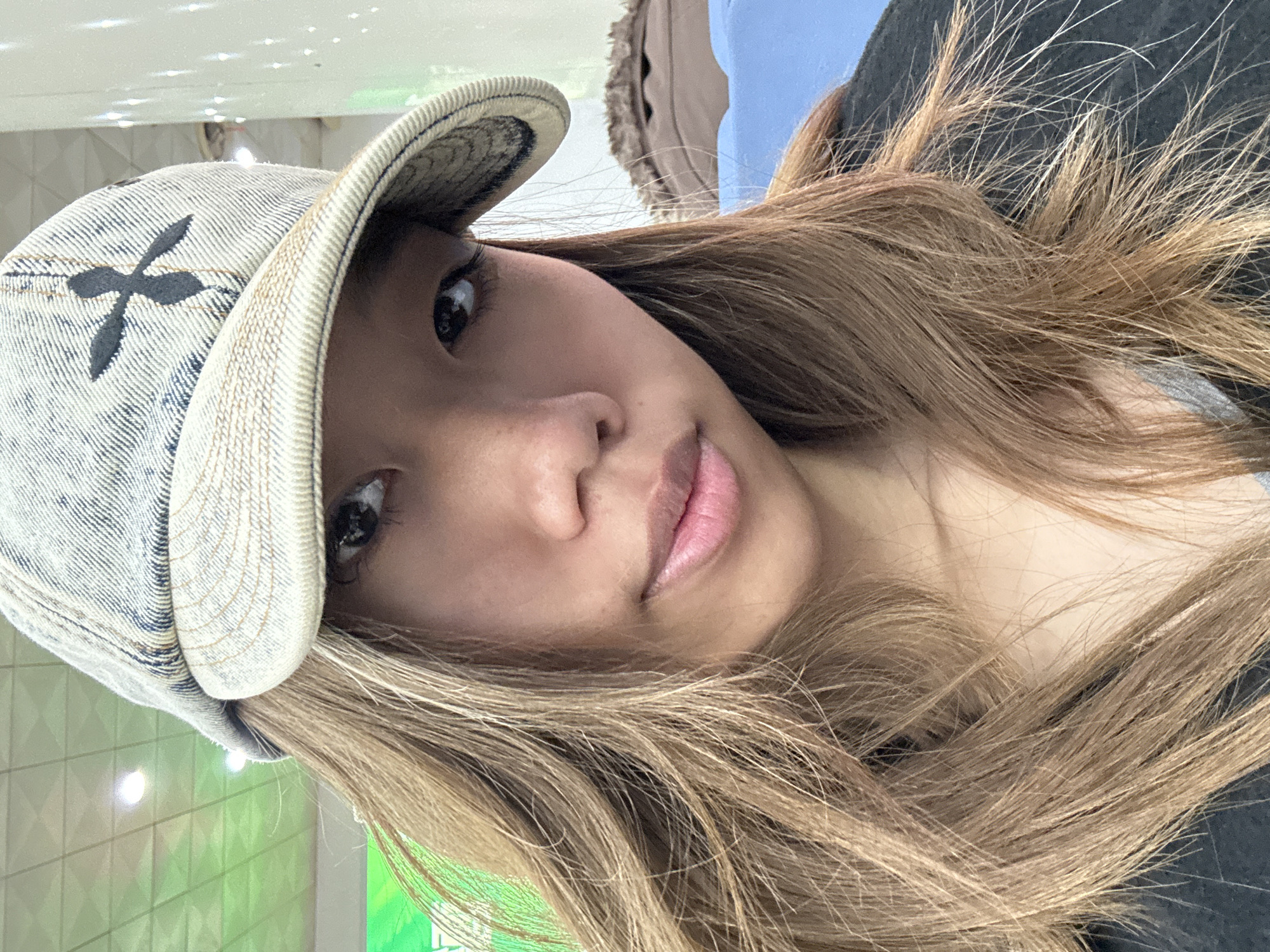 The image is a selfie of a young woman wearing a denim cap with a dark "X" symbol on the side. She has long, light brown hair styled in loose waves. Her skin tone is fair. She appears to be indoors under artificial lighting. It is not possible to determine the exact location, including Lanzhou, China, from this image.