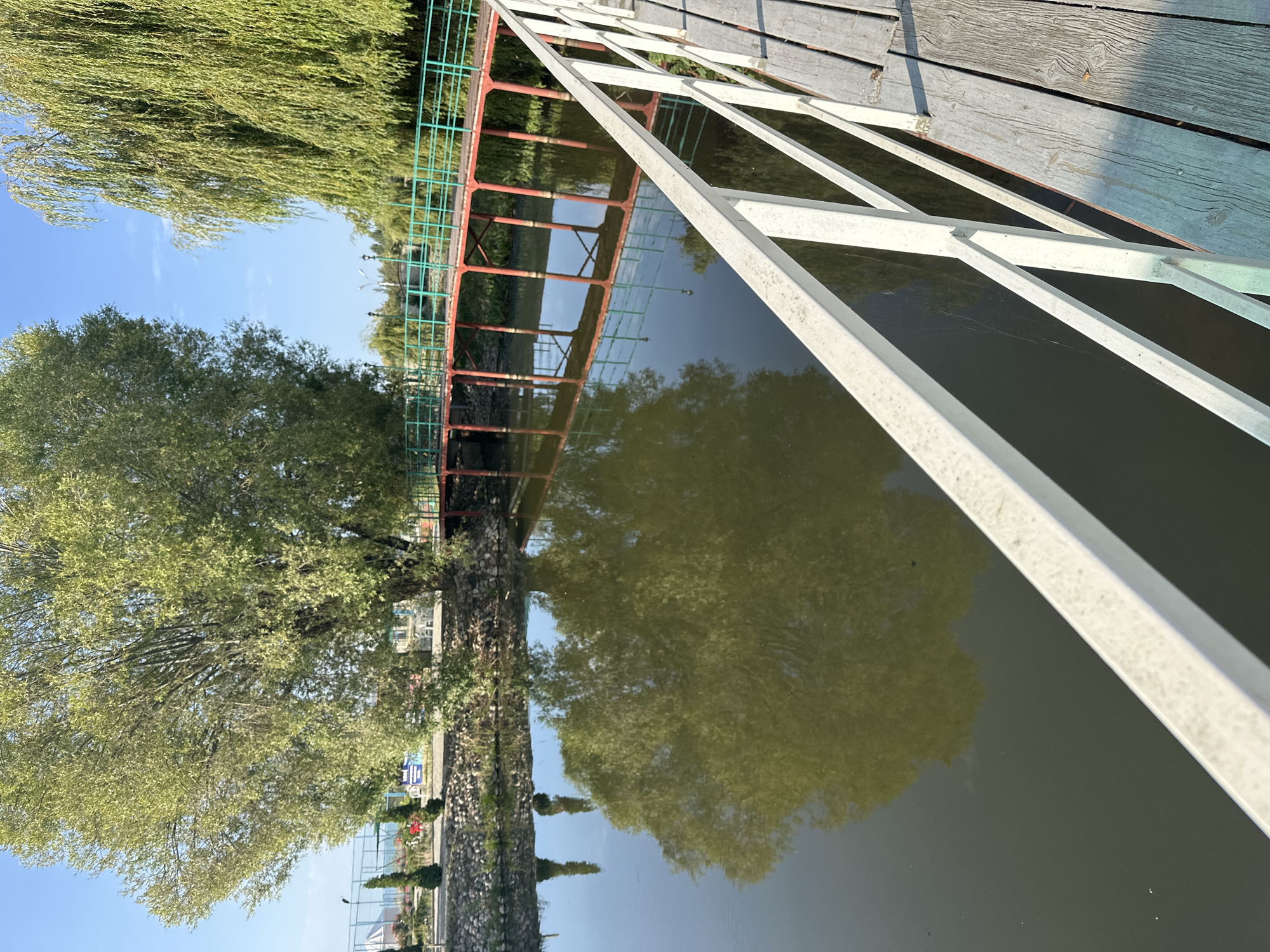 The image shows a tranquil scene in the city of Grigorevka, Kyrgyzstan. A wooden footbridge with white railings extends into the foreground, leading towards a picturesque body of water. On the water's surface, a clear reflection of a large, leafy tree is visible, its branches creating intricate patterns in the still water. In the background, a vibrant red and green bridge spans across the water, supported by sturdy red pillars. Lush green trees, including a weeping willow, flank the scene, adding to the natural beauty of the location. The sky above is a clear, bright blue, suggesting a pleasant day. The overall impression is one of serene natural beauty, characteristic of Grigorevka's landscape.