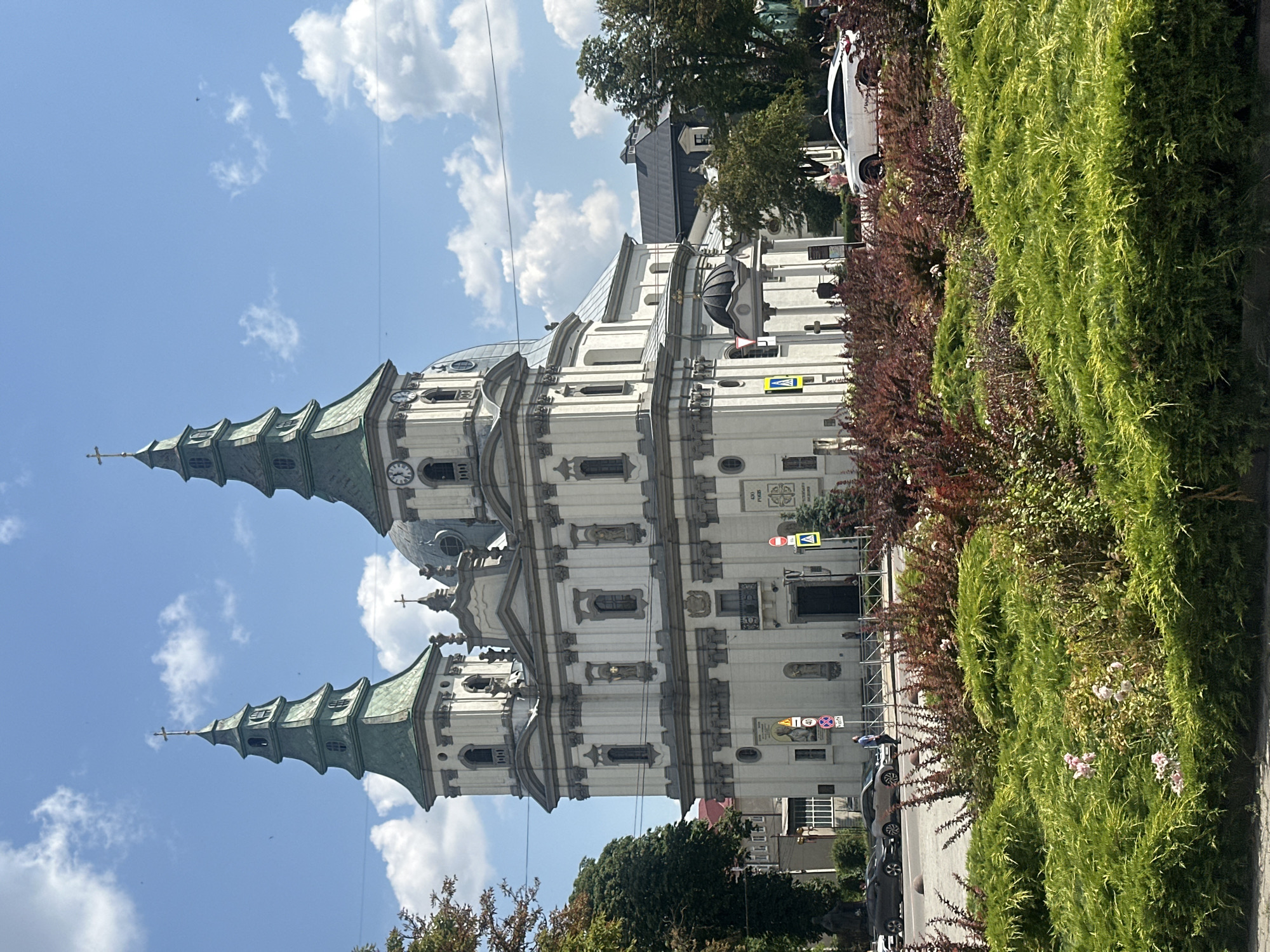 The image shows a large, ornate church with two prominent green-roofed towers, a dome, and intricate white stonework. A clock is visible on one of the towers. The church is situated in a city, with a street and cars in the foreground. There are also decorative bushes and trees in front of the church. The sky is blue with some fluffy white clouds. The building is the Ternopil Castle Church of the Immaculate Conception of the Blessed Virgin Mary in Ternopil, Ukraine.