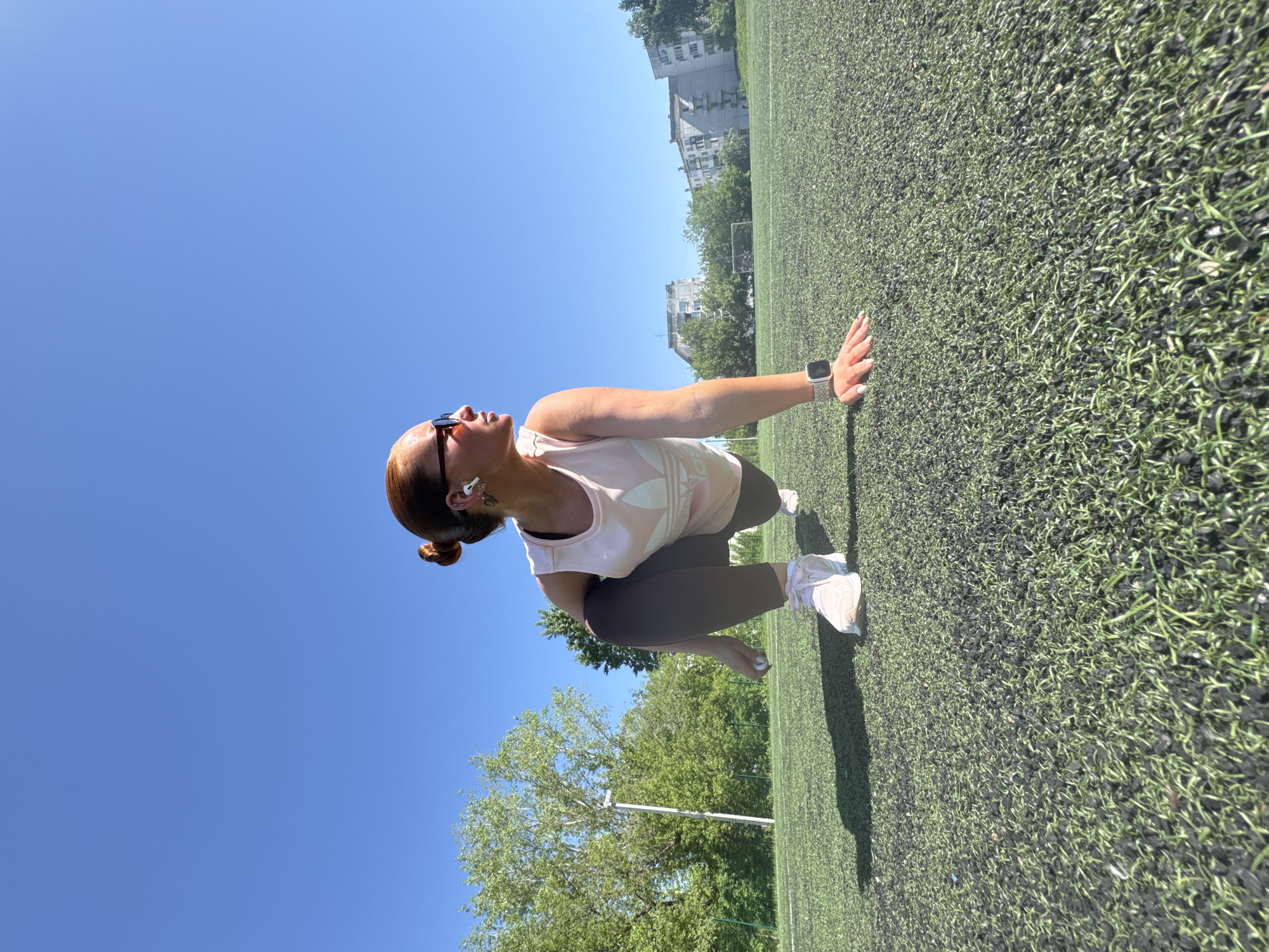 A woman with auburn hair pulled into a bun is performing a dynamic stretching exercise on a green artificial turf field. She is wearing sunglasses, wireless earbuds, a light pink tank top with a visible logo, and dark exercise pants. She has a fitness watch on her wrist. She is in a lunge position with her forward hand touching the turf. In the background, there is a clear blue sky and some trees. On the edge of the field, there are buildings. The location is identified as Chaikovsky, Russia.