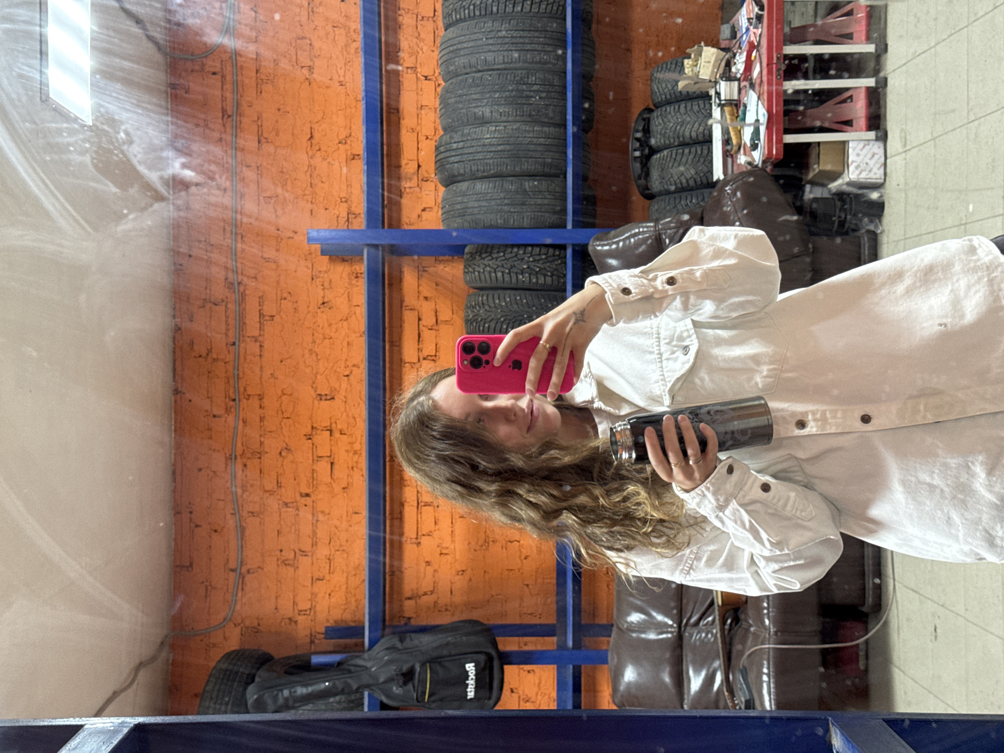 A young woman is reflected in a mirror, holding a pink smartphone in her right hand and a dark-colored tumbler in her left. She has long, wavy hair and wears a light-colored button-down shirt. Behind her is a wall with an orange painted brick pattern, a blue metal shelving unit, and stacks of tires. A black bag rests on the bottom shelf of the unit. Further back, there are brown leather chairs and various garage items. The scene appears to be within a tire shop or garage. There are no signs to suggest this photograph was taken in the city of Chaikovsky, Russia.