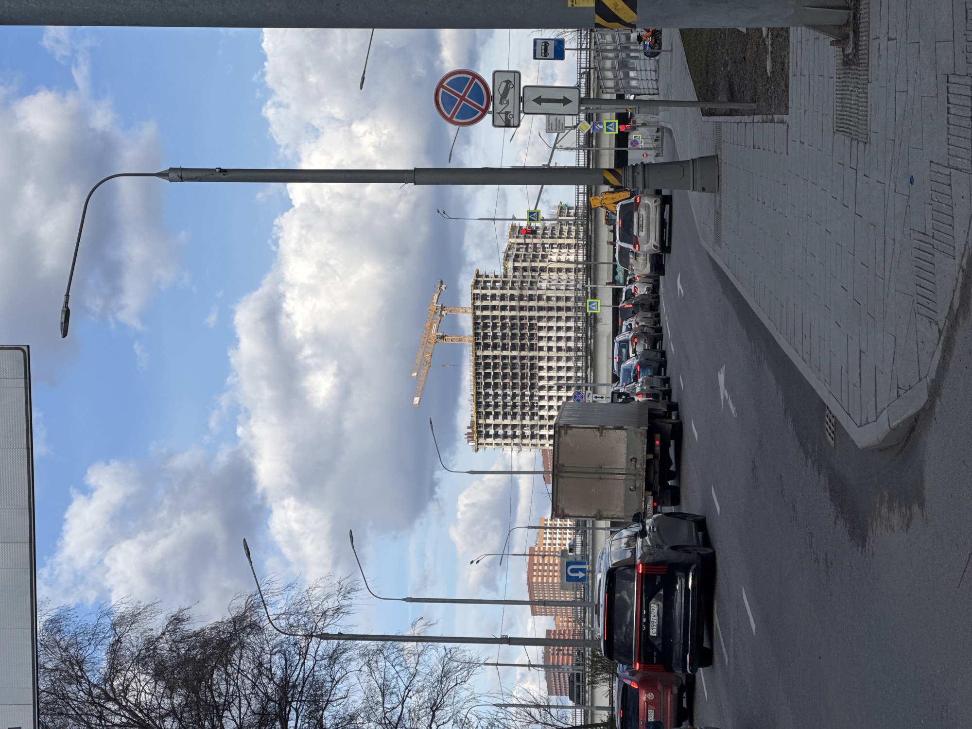 The image depicts a street scene in Moscow, Russia, showing a traffic jam on a road lined with sidewalks and various street furniture. The sky is partially cloudy. A tall building under construction with a yellow crane is visible in the background. Prominent road signs indicate traffic regulations, including "No Stopping" and directions for bus stops and pedestrian crossings. A mix of vehicles, including cars and a large truck, are stalled in traffic. Bare trees are also present in the foreground, suggesting it might be late fall or early spring. License plates are visible on the vehicles, indicating they are Russian. The overall impression is a typical urban scene in a large city with ongoing construction and transportation infrastructure.